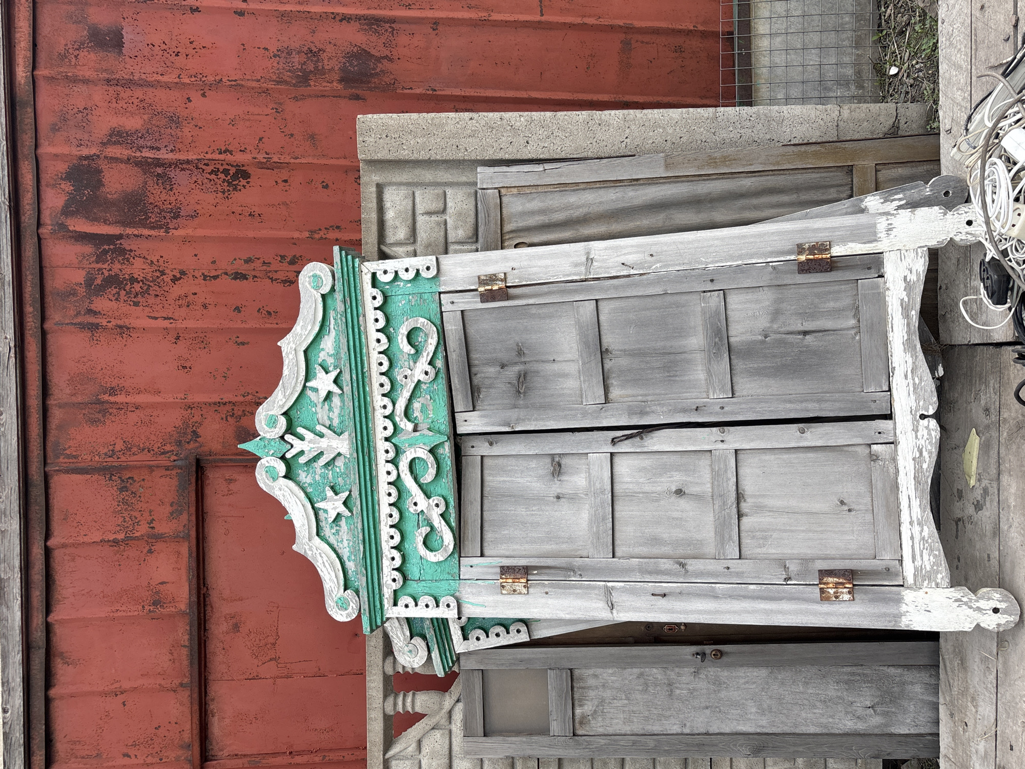 The image shows a collection of weathered architectural salvage items leaning against a rusty red corrugated metal wall, likely in an outdoor storage area in Солонцы, Russia. The most prominent piece is a decorative, turquoise and white wooden window pediment with ornate scrollwork and star motifs. Adjacent to the pediment are various gray wooden shutters or panels, some featuring rusty hinges. A gray concrete or stone slab lies between the pediment and shutters. In the background, wire fencing is partially visible and there is a jumble of white cords. The ground is dirty and rough.