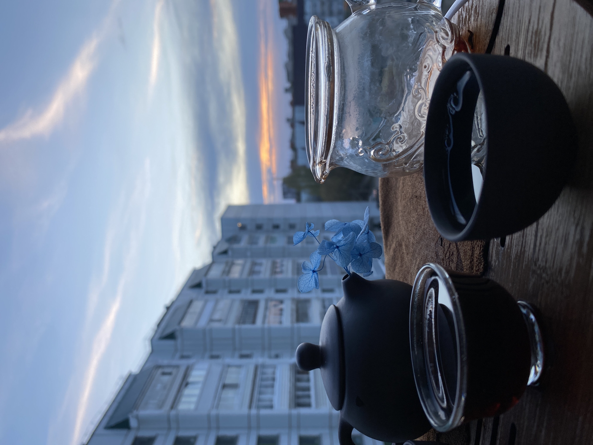 The image depicts a still life composition of teaware set against a backdrop of an urban landscape. A black teapot with blue hydrangeas inside and a clear glass teapot are placed to the right of the frame. Two dark tea cups, one clear and one dark, are near the pots. In the background, a tall, white-walled building with numerous windows is visible, suggestive of an apartment complex. The sky above is a mix of light blue and streaked with white clouds, indicating either dusk or dawn. The image might be captured from a balcony or windowsill. Based on the urban architecture, the location could be Chaikovsky, Russia.