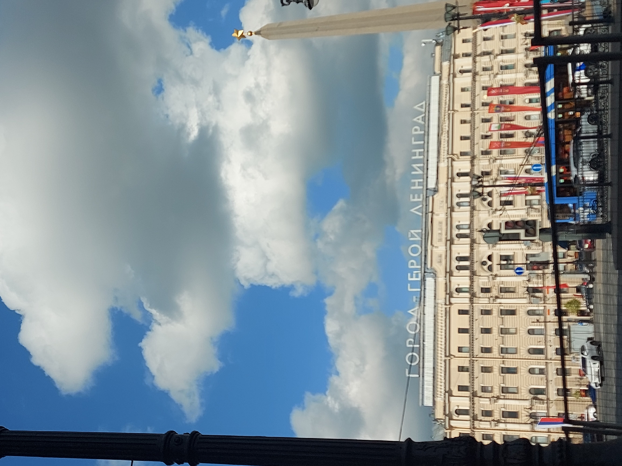 The image shows a view in Saint Petersburg, Russia, with a partially obscured view of the Hero City Obelisk and a multi-story building decorated with red banners. The sky is visible with a mix of blue sky and white clouds. A dark, ornate metal structure is present in the foreground, likely part of a fence or lamp post. The text "ГОРОД-ГЕРОЙ ЛЕНИНГРАД" (Hero City Leningrad) is visible on the building's facade. A blue and white bus and several cars are on the street in front of the building.