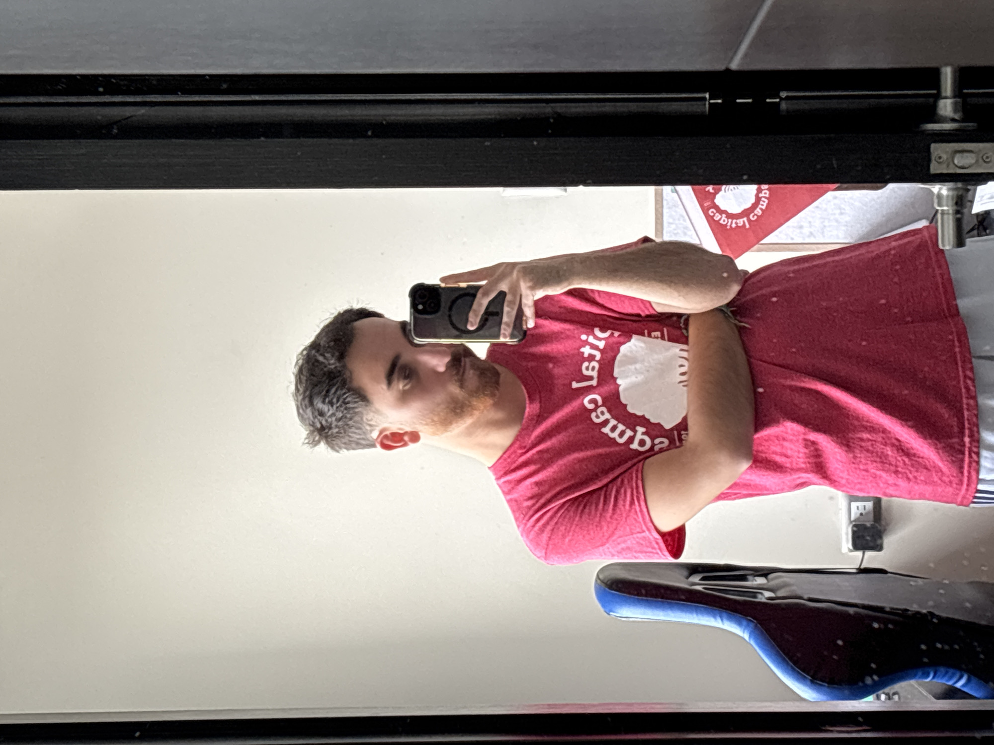 A man is taking a selfie in a bathroom mirror. He is wearing a red t-shirt. Behind him is a red and white rug. A bass guitar in a dark case with blue trim rests against the wall. There's an electrical outlet with a black device plugged into it. The mirror is framed by a black cabinet. The location cannot be determined from the image.