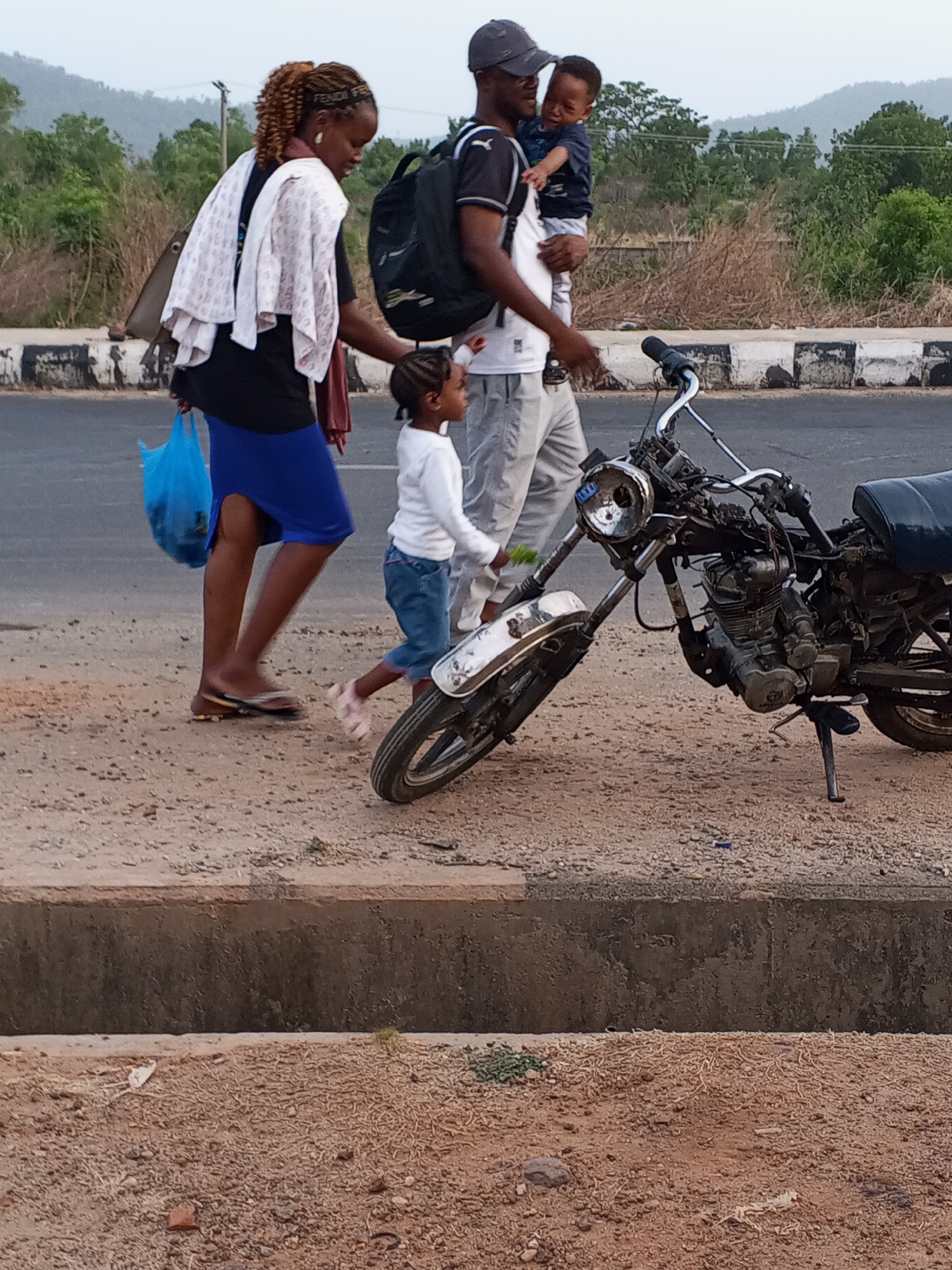 On a dirt and gravel shoulder adjacent to a two-lane road in Lokoja, Nigeria, a family walks alongside a disassembled motorcycle propped on its kickstand. A man, wearing a cap, dark t-shirt, light pants, and a backpack, carries a young child. A woman, wearing a dark t-shirt, blue skirt, and flip-flops, holds a blue plastic bag and has a white towel draped over her shoulder. A young girl, wearing a white long-sleeved shirt and denim shorts, walks beside the woman. The motorcycle is stripped down, exposing the engine and frame. Trees and distant hills form the background.