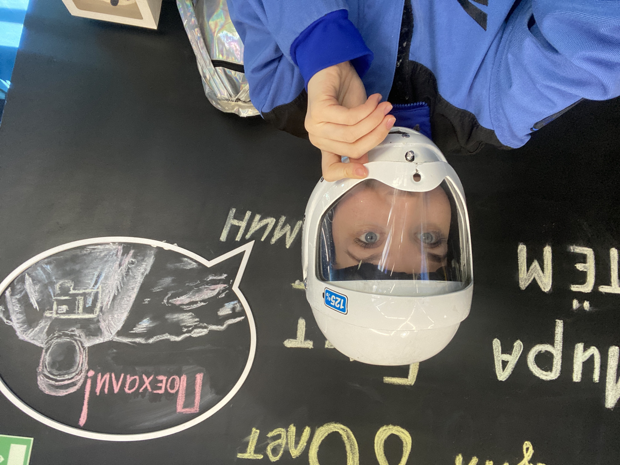 A person wearing a blue jacket and a white helmet with a clear visor is positioned above a black surface covered in white and yellow chalk writing. The helmet obscures the person's face, but their eyes are visible through the visor. A drawing of an astronaut in a thought bubble and text is on the left. The scene is likely indoors, based on the artificial lighting. The phrase "Поехали!" and other Russian words are present, suggesting the location is Izhevsk, Russia.