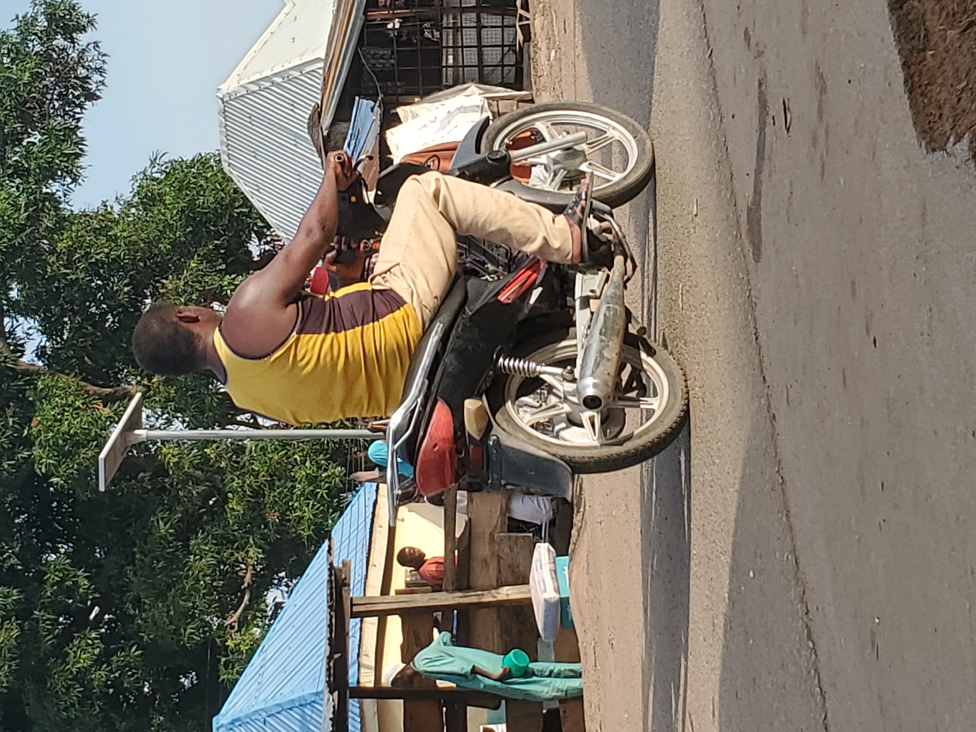 A man wearing a yellow tank top and tan pants is sitting on a black motorcycle in Mararaba, Nigeria. The motorcycle is parked on a paved road. In the background, there are trees, buildings with corrugated metal roofs, and a small wooden structure with a blue tarp.
