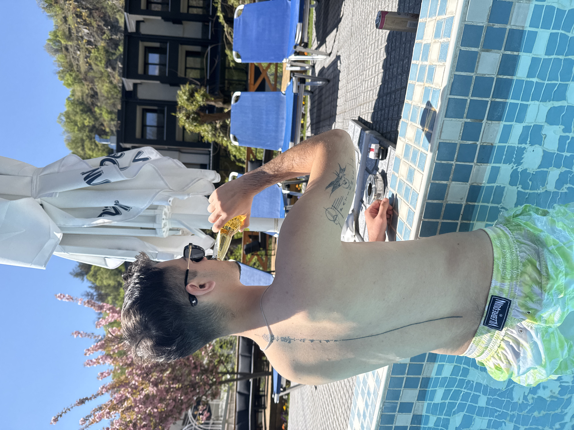 A fair-skinned individual stands partially submerged in a pool, positioned in front of the tiled edge of a swimming pool. The person has tattoos on their arm and back, and they wear dark sunglasses. They are drinking from a glass bottle. They are wearing green and white swim trunks with a logo on the side.
Behind the individual, there's an umbrella, blue lounge chairs, and a building with multiple windows. A can is on the tiling next to the swimming pool. A section of brick pavement is also visible. Pink flowers bloom from a tree to the left of the pool.
The setting suggests a warm, outdoor environment. Based on additional context, it is inferred that the location is Marchevo, Bulgaria.