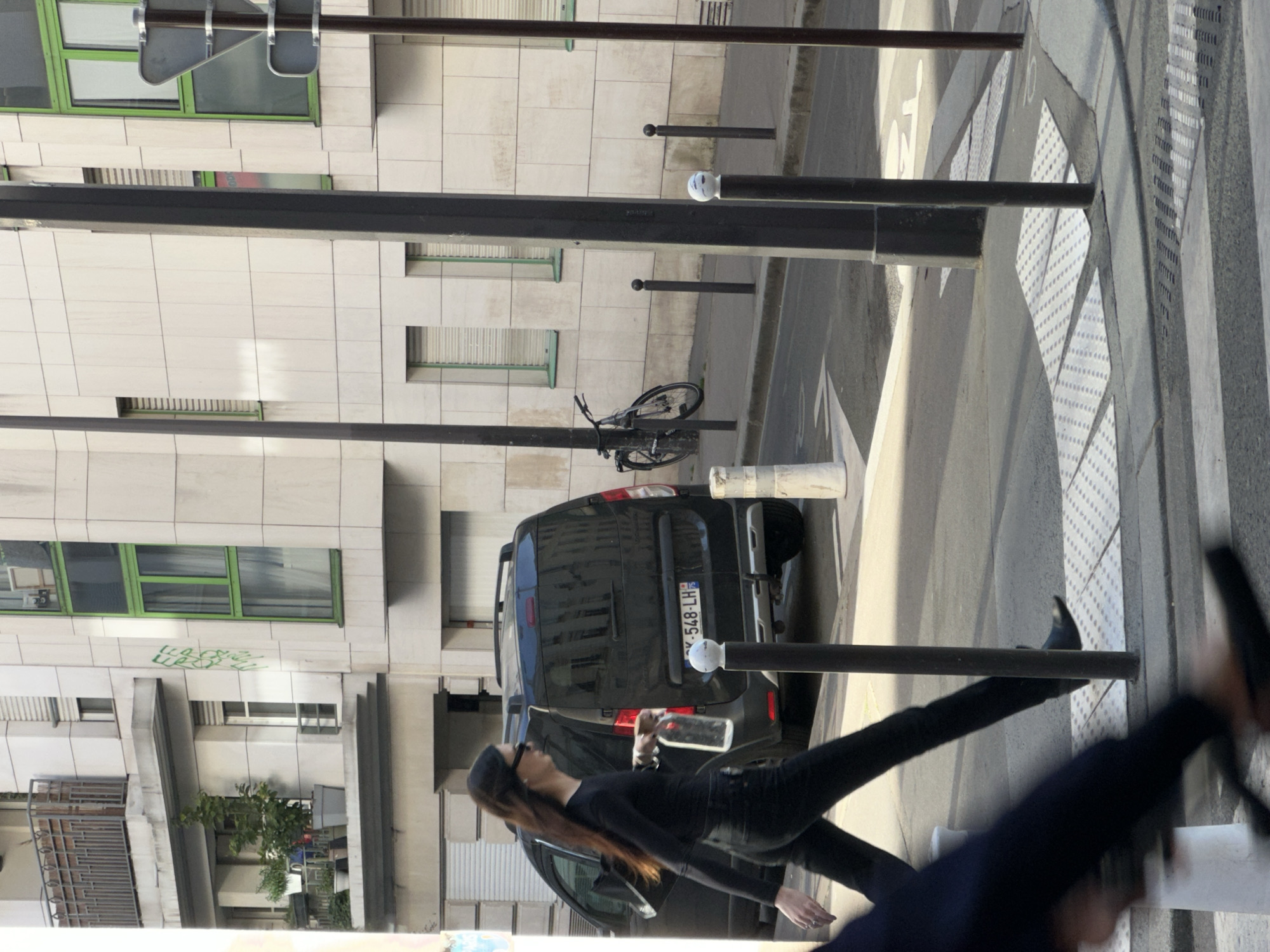 The image depicts an urban street scene in Paris, France, captured in bright daylight. The perspective is slightly elevated, looking down a street alongside a modern building and a road.
In the foreground and mid-ground, a woman with long brown hair, wearing a black long-sleeved top, black pants, and sunglasses (or a cap), is walking on a paved sidewalk. She is holding a clear plastic bottle in her right hand, which is extended. Her posture suggests she is in motion. A partial, blurred view of another person, likely walking in the same direction or towards the camera, is visible in the bottom right corner.
To the left, a multi-story building with a light-colored, tiled facade dominates the background. It features several rectangular windows, some framed in a distinct green, others with external roller blinds drawn down. Faint, illegible green graffiti is visible on one section of the building's facade.
On the street, a black van is parked, facing away from the viewer. Its French license plate "X-548-LH" is clearly visible. A bicycle is chained to a black pole near the van. Several other black street bollards line the edge of the sidewalk. The road surface includes a clear zebra crosswalk with patterned tactile paving for the visually impaired. Faint white road markings, including fragments that look like "TO" and the number "10," are also present.
The scene is well-lit by natural sunlight, indicating a clear, possibly sunny, day. Shadows are visible on the ground and buildings. The architecture and vehicle license plate confirm the location as urban France.