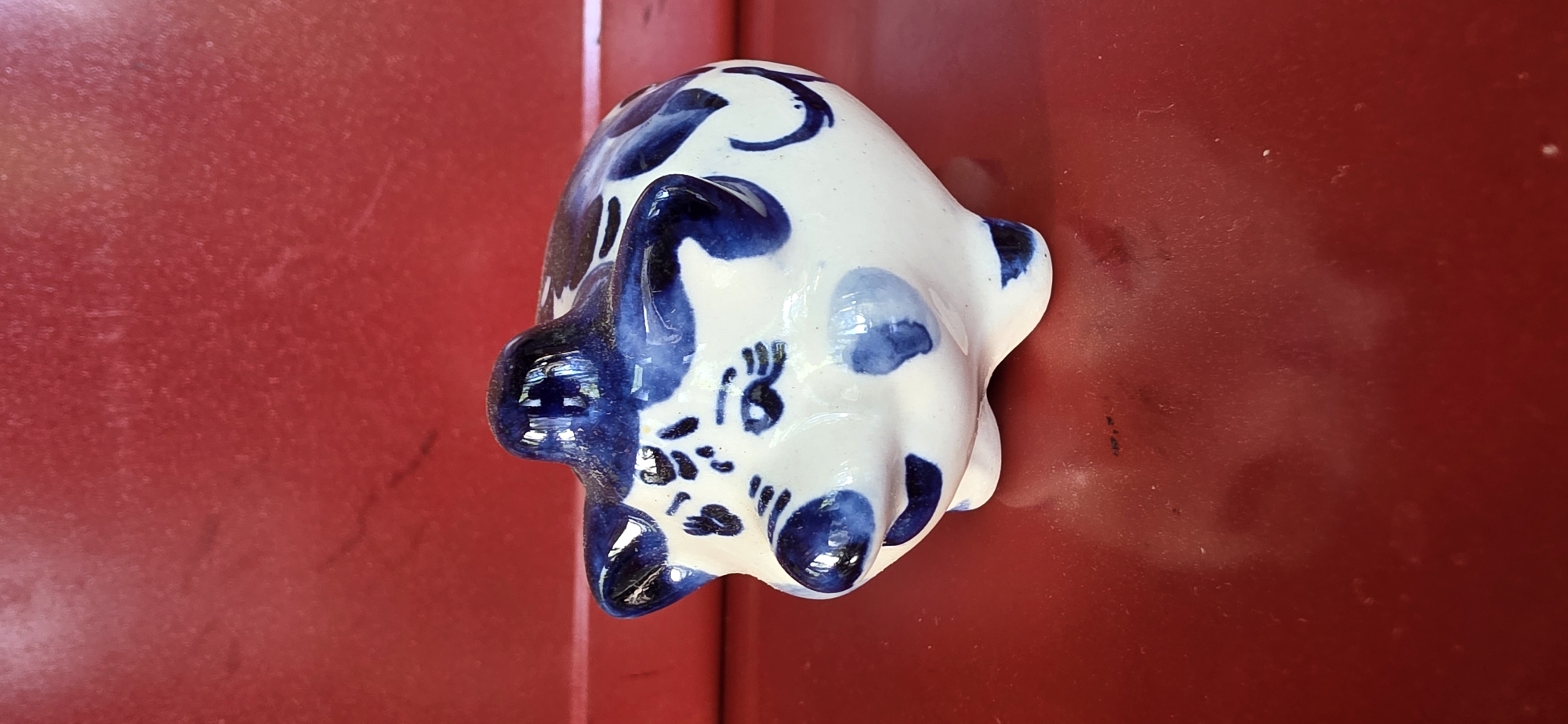 A ceramic pig figurine, painted with blue floral patterns on a white background, sits on a shiny red surface. The figurine is facing left, with its snout slightly elevated. The background is a solid red color with some subtle texture. The lighting casts a reflection of the figurine onto the red surface below. The scene appears to be indoors, given the lighting and the smooth, uniform background. The location is not specified in the image itself.