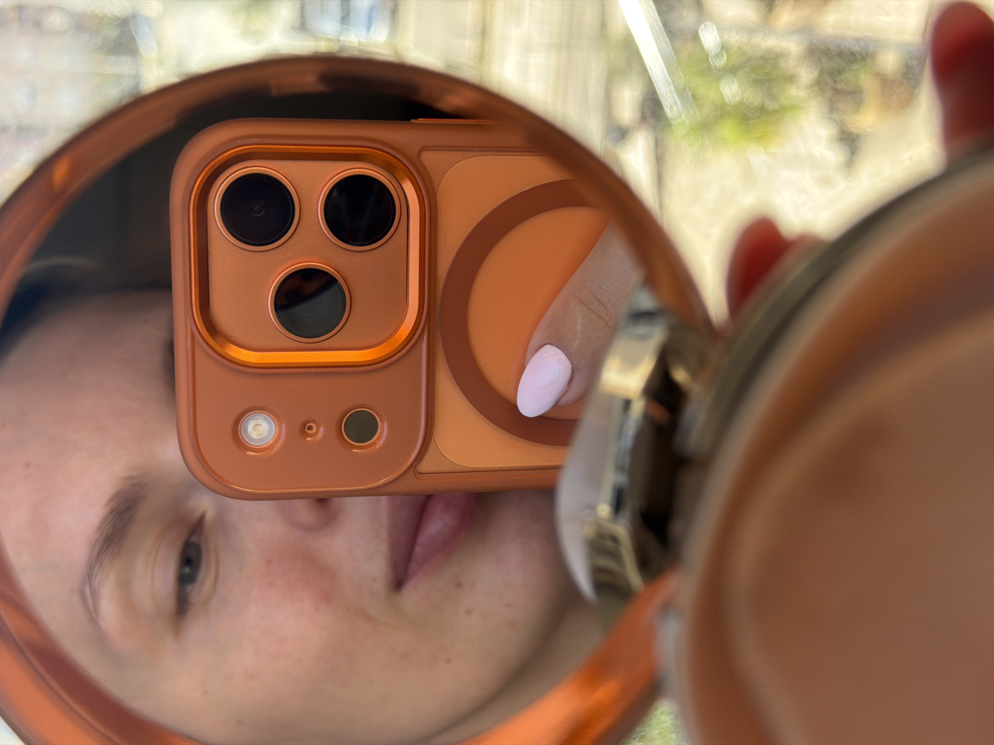 The image is a close-up, reflective shot captured in a round mirror with an orange or copper-toned frame. The primary subject in the reflection is a person's face, partially visible, including an eye, eyebrow, nose, and mouth, all framed by the mirror's edge. Their expression is neutral.
Dominating the reflection is the back of an orange smartphone, presumably an iPhone given its distinct triple-lens camera module, which includes three prominent black camera lenses, a flash, and a smaller sensor. The phone is encased in a matching orange protective cover that features a slightly raised border around the camera array and a subtle circular design element on its back. A finger, adorned with light pink nail polish, is seen holding the phone, with part of the hand visible on the right side of the reflection.
The background of the reflection is softly blurred, showing a mix of light green and yellow tones, which suggests an outdoor scene like foliage or distant buildings, viewed through a window or in an outdoor setting, likely during the daytime. The act of taking a mirror selfie or a reflection photograph is the central activity. No text is visible in the image. The scene is situated in Budva, Montenegro.