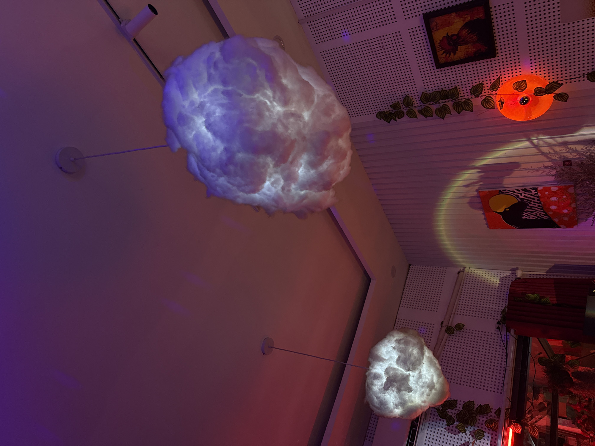 The image displays an artistically lit indoor space, likely a cafe or bar, characterized by whimsical decor and ambient lighting, captured in the evening or at night. The central focus includes two suspended light fixtures resembling glowing white and purplish clouds. The larger cloud lamp hangs prominently from a smooth, light-colored ceiling section in the upper left, while a smaller one is visible in the lower right, also emitting a soft glow.
The ceiling features a combination of smooth surfaces and perforated acoustic panels. Various decorative elements are arranged across these surfaces. These include a small framed artwork depicting a colorful rooster or bird in red, black, and yellow tones. Adjacent to this, an orange circular light, possibly a stylized sun or fruit, is entwined with artificial green vine-like foliage. Below this, another abstract artwork or poster in black, white, red, and yellow can be seen. Further artificial foliage is draped across the perforated panels and around structural elements. Red and orange neon lights or strips are visible on the right side of the frame, contributing to the vibrant, artificial illumination.
The overall lighting consists of deep purple, pink, and orange hues, casting a warm and inviting glow across the room. No people or specific activities are visible, emphasizing the decorative and atmospheric elements of the space. There is no discernible text within the image. The setting appears to be a creatively designed interior in Đà Nẵng, Vietnam, intended for a relaxed or social ambiance.