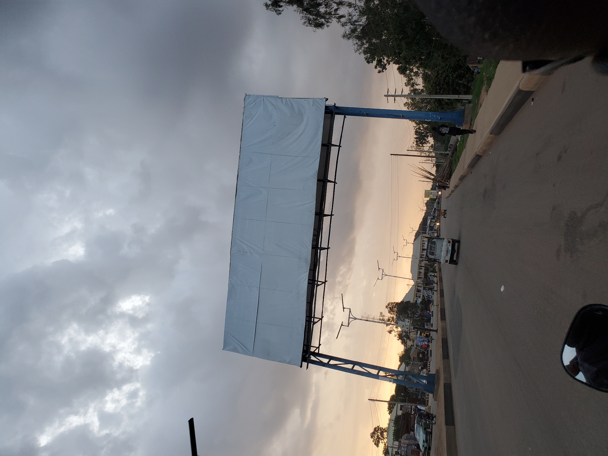 The image captures a street scene in Gura Top, Nigeria, at dusk. A large, empty billboard dominates the upper center of the frame, supported by a blue metal structure that spans across the road. The sky is filled with heavy, grey clouds, with some patches of lighter sky suggesting the setting sun. A road stretches into the distance, with a few vehicles visible, including a white pickup truck and a car. Pedestrians are present on the sidewalk, with one individual walking along the side of the road. The environment appears to be a mixed-use area with buildings lining the street and trees on the right side of the frame. Power lines and streetlights are also visible. The perspective of the shot is from a low angle, possibly from a vehicle.