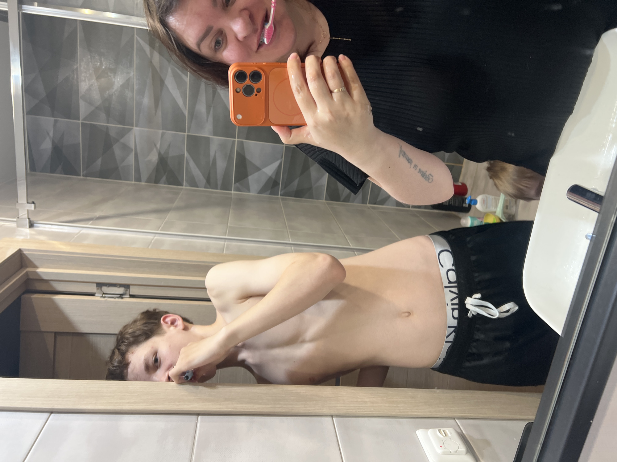 A photograph shows a reflection in a bathroom mirror. Two people are visible in the mirror. One person, presumably female, is holding a cell phone in an orange case while brushing their teeth. A second person, with a shirtless torso and Calvin Klein shorts, is also brushing their teeth. The bathroom features patterned tile walls and a white sink. The environment includes toiletries and a light switch on the wall. There is no information in the image to determine if the location is in the city of Noviy, Russia.