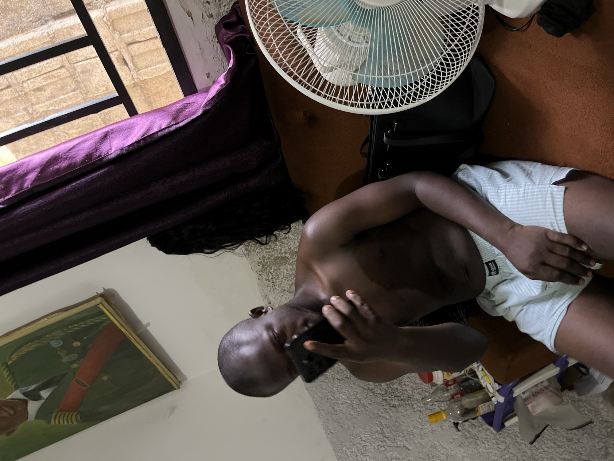 A shirtless man is seated on the floor talking on a mobile phone. He wears striped shorts. A white fan sits on a black bag behind him. A window with purple curtains is visible in the upper left. An unframed portrait is displayed on the wall. The location is in a room. The city and country are Abuja, Nigeria.
