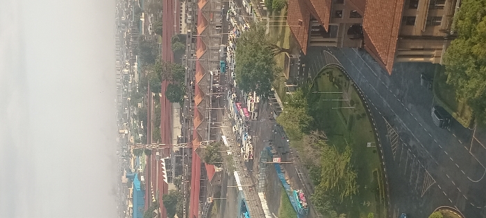 An aerial view shows a city street congested with various vehicles, including buses, flanked by buildings with reddish-brown roofs. A prominent structure with similar roofing is positioned to the right, accompanied by a curving road and greenery. In the distance, a hazy atmosphere obscures the cityscape. A construction crane is visible amidst the buildings, suggesting ongoing development.