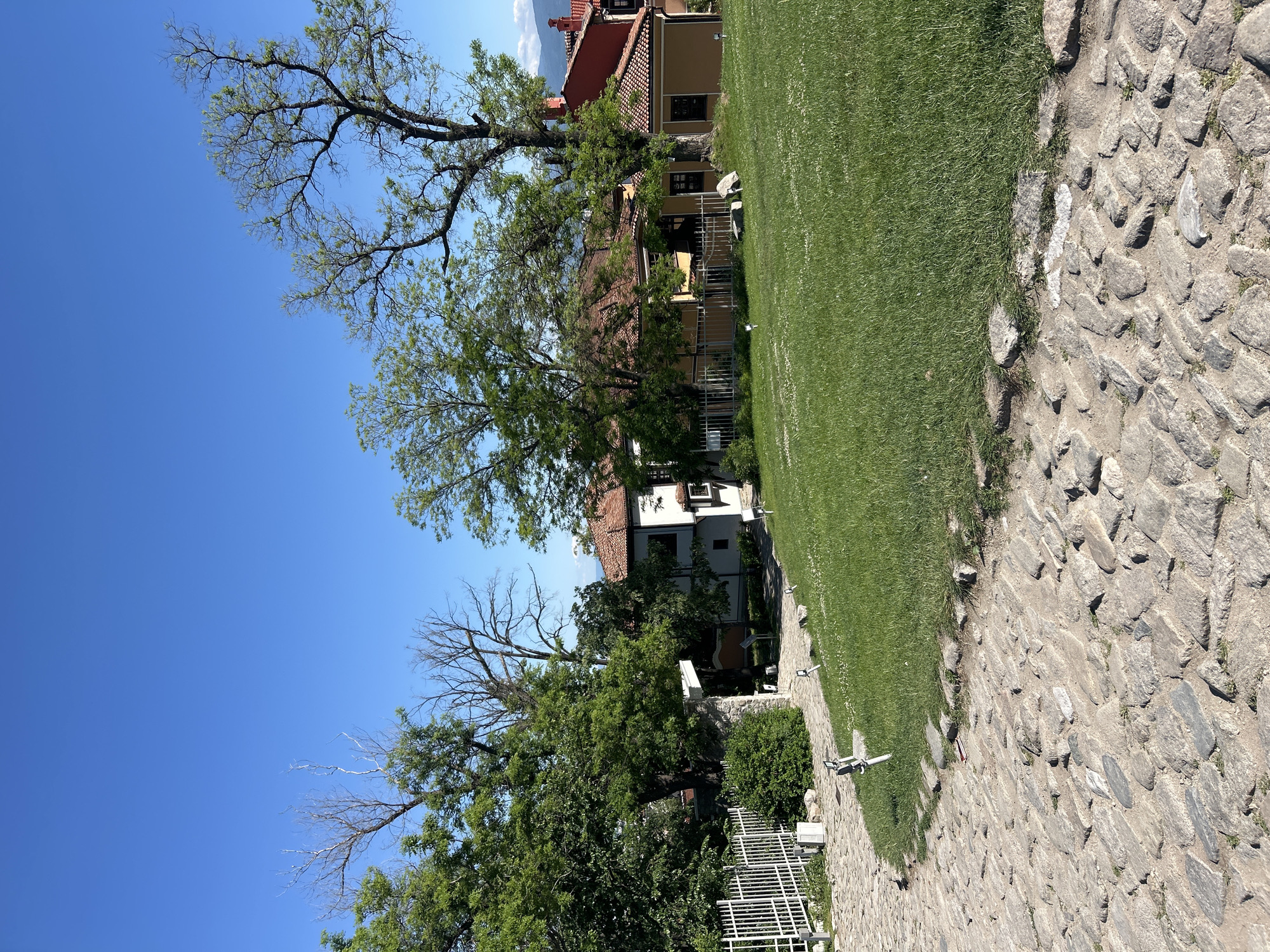 The image captures a scene in Plovdiv, Bulgaria, featuring a stone-paved pathway alongside a grassy area. Several buildings with red-tiled roofs are partially visible behind trees. The sky is clear blue. A white fence is seen in the lower left, and there are trees with varying degrees of foliage throughout the frame. Lighting fixtures are attached to the low retaining wall separating the grass and the paving.