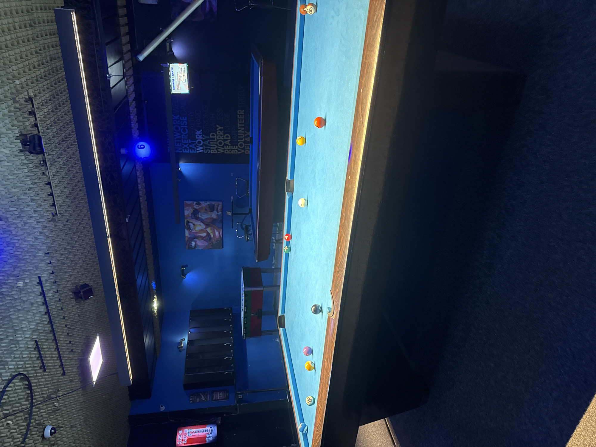 The image shows a pool hall with blue walls and blue felt pool tables. Several billiard balls are scattered across one of the tables. There is also a foosball table visible in the background, along with some lockers. The lighting is dim, with some blue and white lights creating a moody atmosphere. A large graphic displaying motivational text such as "NETWORK WELL", "EXERCISE DAILY", "EAT HEALTHY", and "WORK HARD" is mounted on the back wall. A small screen displaying the word "ВИЛЯРДА / ДАРТС" (Billiard / Darts) is also visible on the back wall. The scene appears to be an indoor recreation area or bar. The location is not specified to be Yambol, Bulgaria, and no people are present in the image.