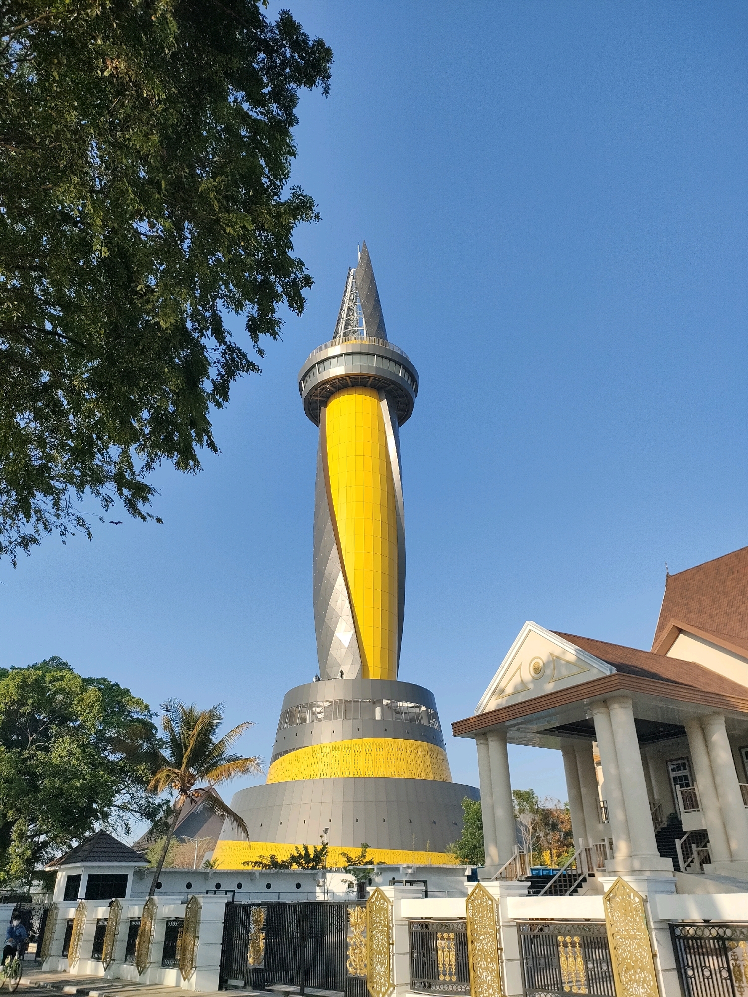 This image captures the Siring Patung Bekantan, a distinctive landmark in Banjarmasin, Indonesia, known for its prominent statue of the proboscis monkey (Bekantan). The central focus is the striking, modern tower with a spiraling design in yellow and gray, topped with a pointed spire. The tower is situated in a landscaped area, with a lush green tree dominating the left side of the frame, its branches reaching towards the clear blue sky. To the right of the tower, a classical-style building with columns and a brown roof is visible, suggesting it is part of the Siring complex or a nearby structure. In the foreground, a white fence with ornate gold details and black metal gates separates the area from a pathway or road. A few people are present; one individual is seen cycling on the pathway, and others are standing near the fence. A palm tree adds a tropical element to the scene, which is bathed in bright sunlight, indicating it is daytime.
