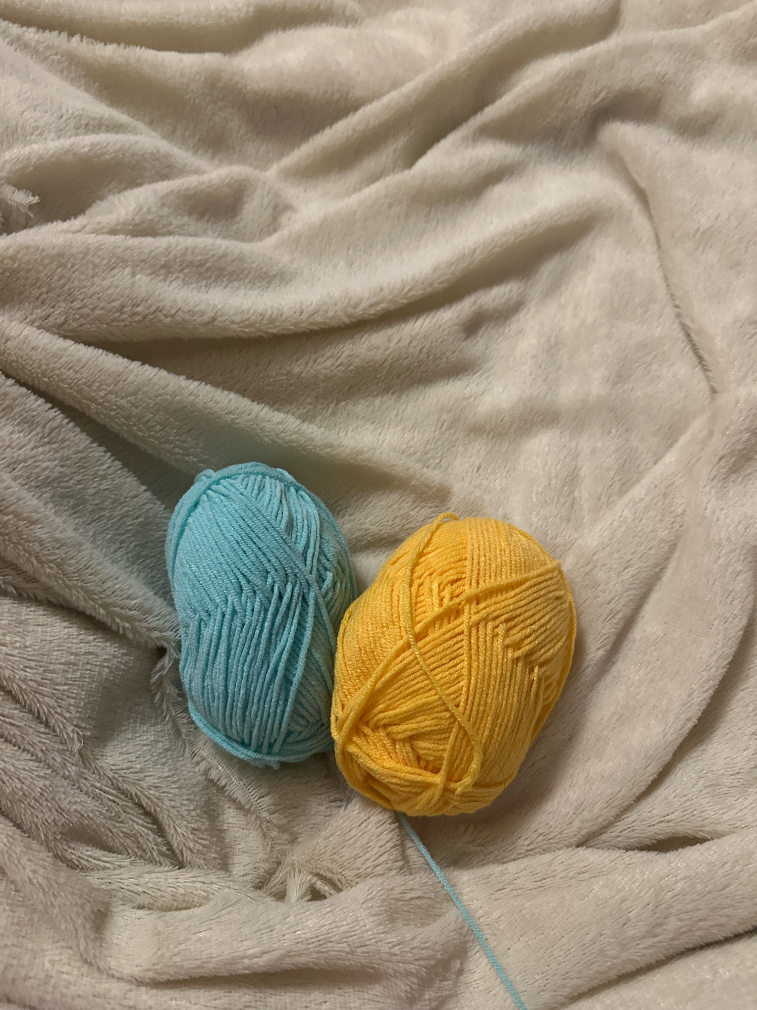 Two balls of yarn, one blue and one yellow, rest on a white, textured blanket. A light blue strand of yarn extends from the yellow ball, trailing across the blanket. The background is soft and blurred, suggesting a shallow depth of field. No specific location in Gyumri, Armenia, can be identified from the image.