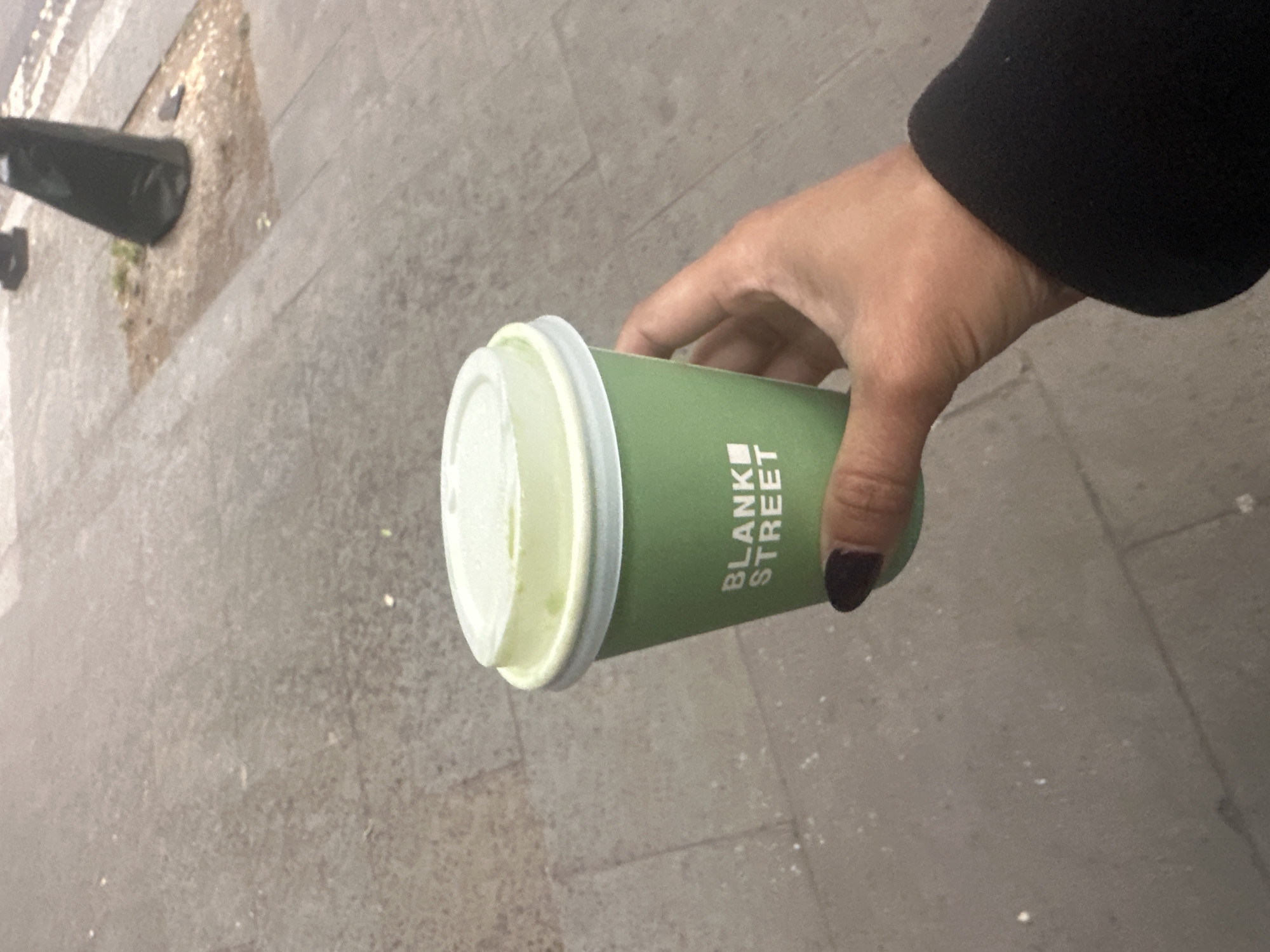 A hand holds a green paper coffee cup with a white plastic lid, branded with "BLANK STREET" in block letters. The hand has dark nail polish. The background is a gray, concrete-paved sidewalk, possibly in a pedestrian area. A black object, perhaps a street fixture or a sign base, is partially visible in the upper left corner. The scene likely occurs on a street in London, United Kingdom, given the "BLANK STREET" coffee chain's prevalence there.