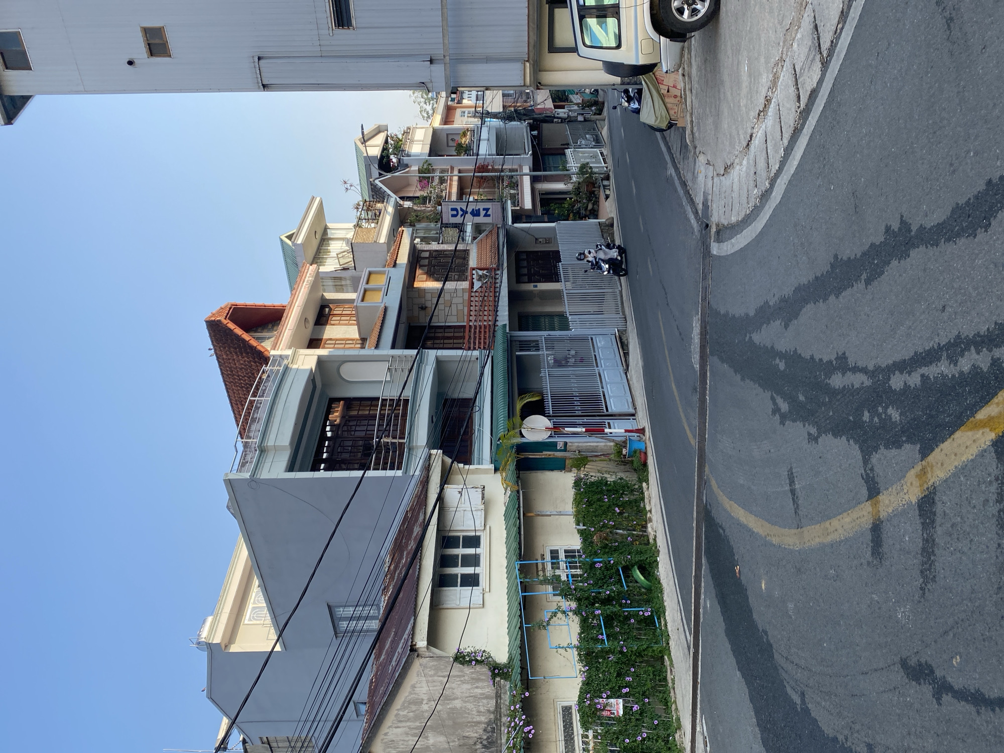The image depicts a street in Da Lat, Vietnam, lined with narrow, multi-story buildings of varying architectural styles. The buildings are close together, with some displaying balconies and roof gardens. A blue sign with the letters "ZKCC" is visible on one of the buildings. A motorcycle is parked on the street. The asphalt street shows signs of wetness, with dark streaks and a faded yellow line. The sky is clear and blue. Electrical wires are visible in the foreground.