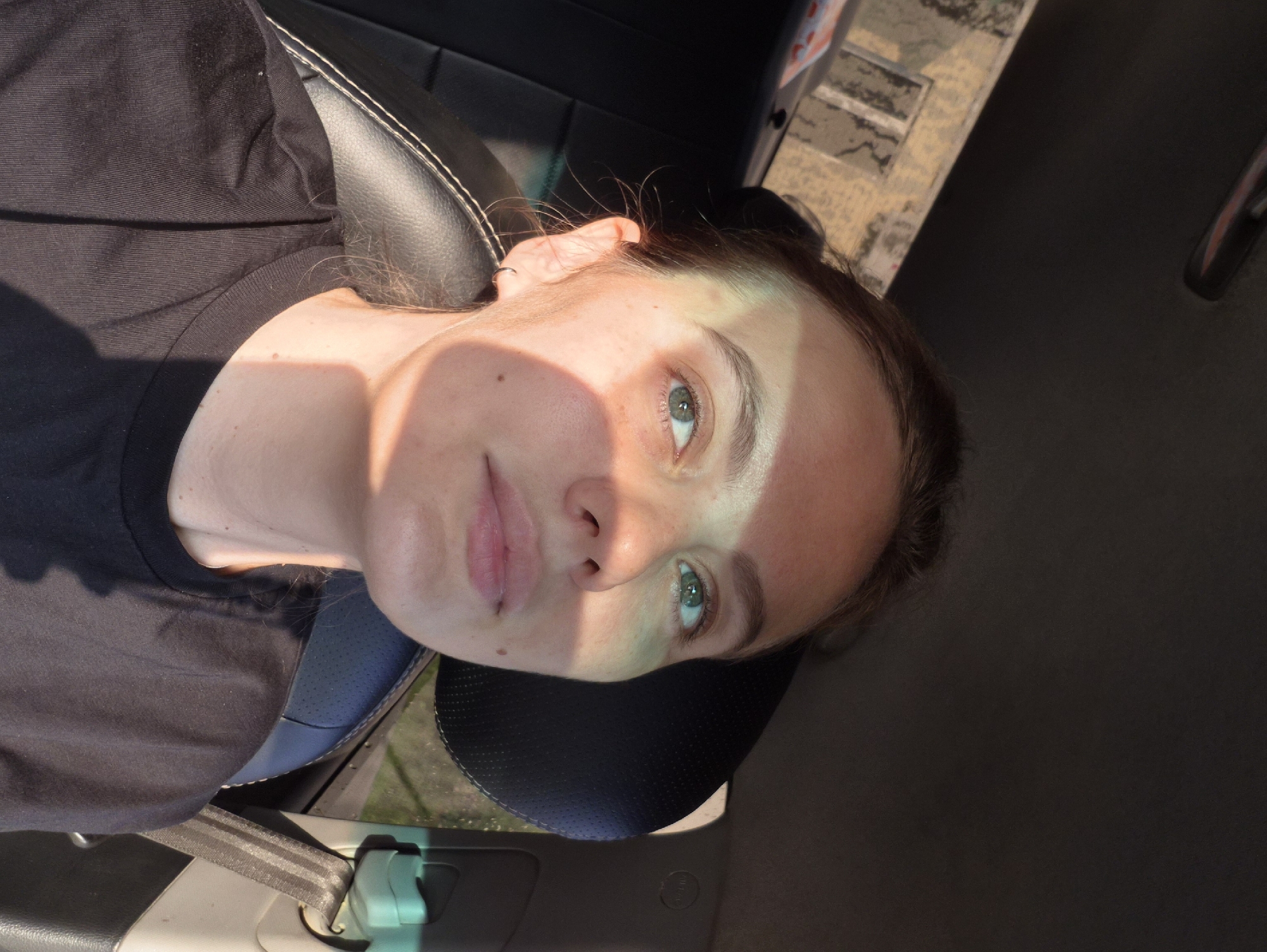 A young woman with light skin and light brown hair is looking up and to her right. She is sitting in a car, her head and shoulders are visible. She is wearing a black t-shirt. A shadow is cast across the left side of her face, obscuring her left eye and forehead. The background of the image shows the interior of a car, including a black leather seat and a portion of the car's headrest and a seatbelt. Outside the car, a building with windows is visible, suggesting an urban environment. The photograph was taken in the city of Chaikovsky, Russia.