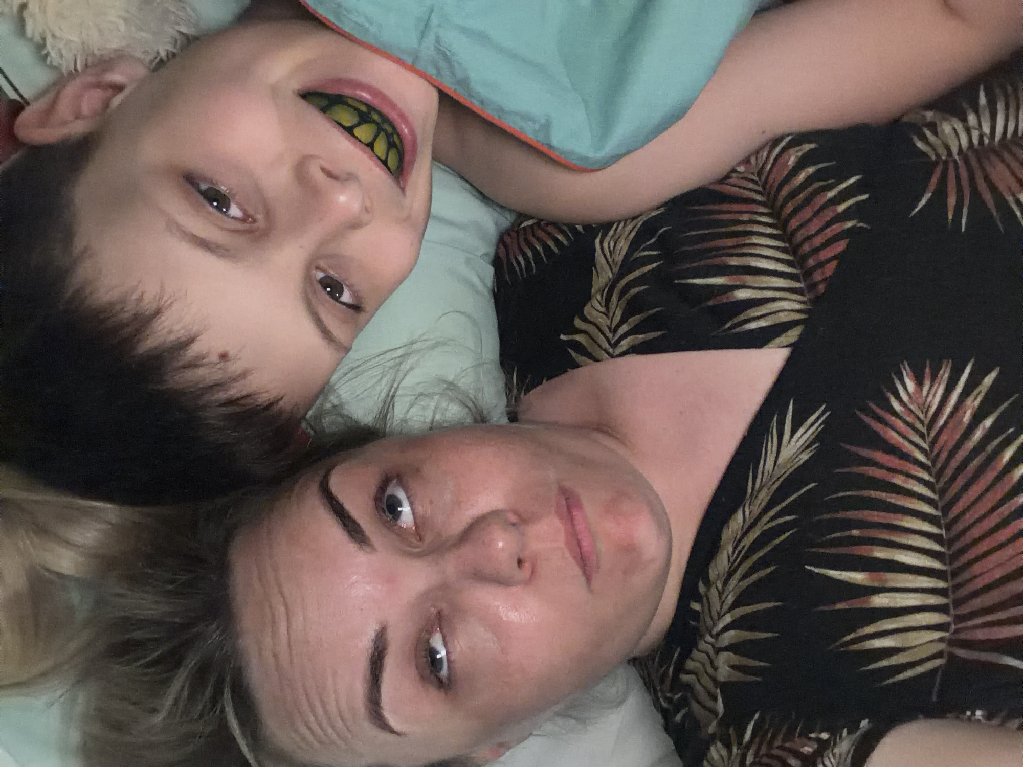 A photo of a woman and a child lying on a bed. The woman has light skin, blonde hair, and is wearing a black shirt with a pattern of golden palm fronds. The child is positioned above her, with dark hair and a yellow-and-black mouthguard in his mouth. They are both looking at the camera. The bed has a light blue sheet, and there is a light-colored, fluffy object at the top left of the frame. The photo is rotated slightly. No information suggests this image was taken in Votkinsk, Russia.