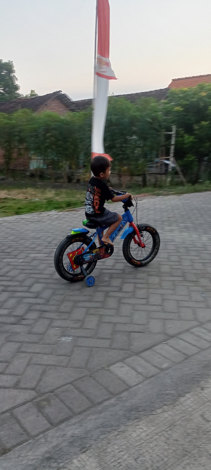A young boy is riding a blue and red bicycle with training wheels on a paved pathway. He is wearing a black t-shirt with a graphic design and dark shorts. The boy is looking to his right, and his bare feet are on the pedals. The bicycle has a green seat and a blue fender. In the background, there is a white and red flag on a pole, green trees, and buildings with red roofs. The scene appears to be outdoors in the town of Gempolklutuk, Indonesia. The ground is covered in interlocking brick pavers.