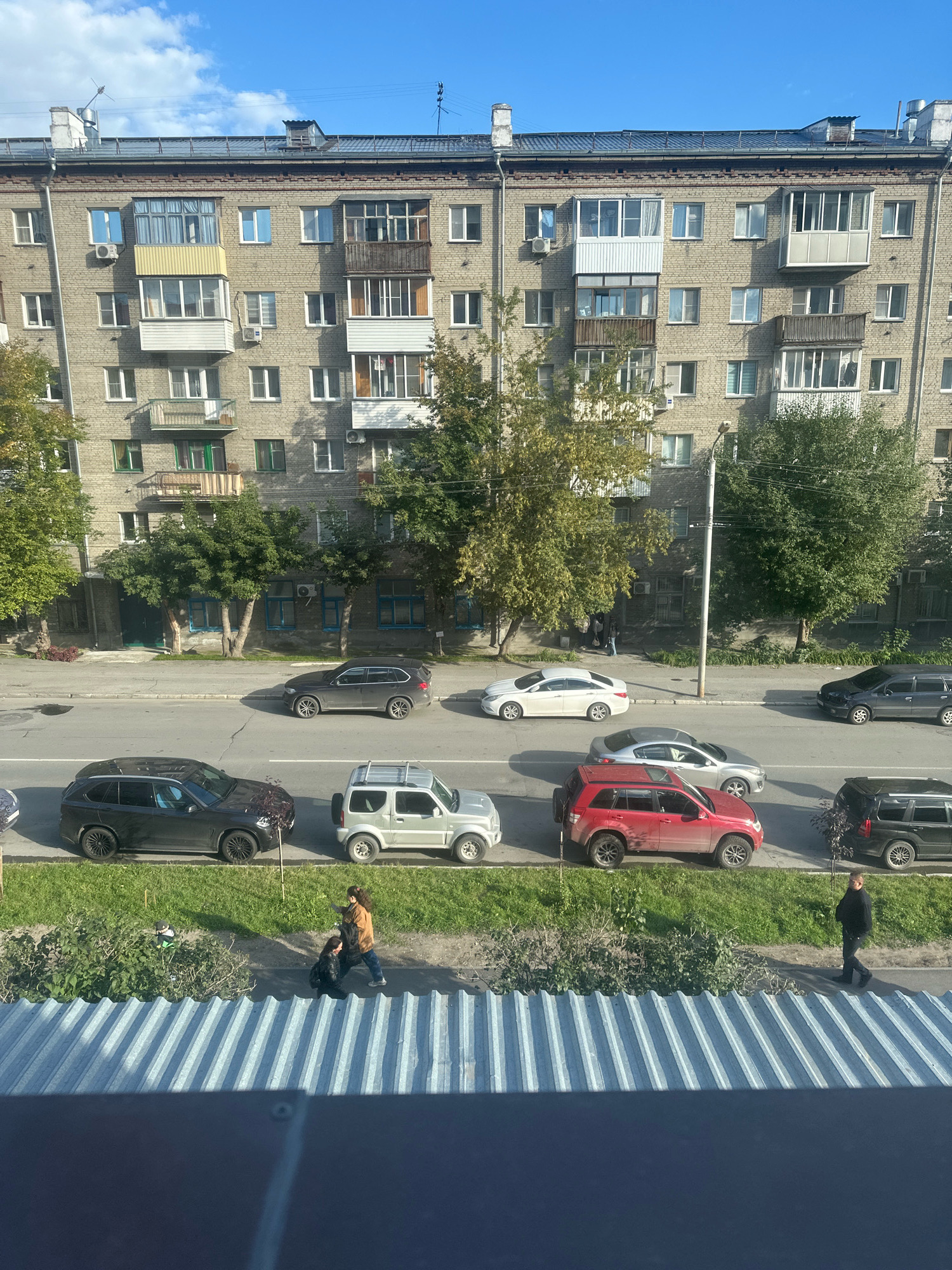The image captures a street scene with a multi-story apartment building in the background. Several cars are parked and driving on the road. In the foreground, there is a sidewalk and a grassy area. Two individuals are visible on the sidewalk, one bending down and the other standing. Another person is walking on the sidewalk further down. The sky is blue with some clouds. The architecture of the building suggests a post-Soviet era construction.
