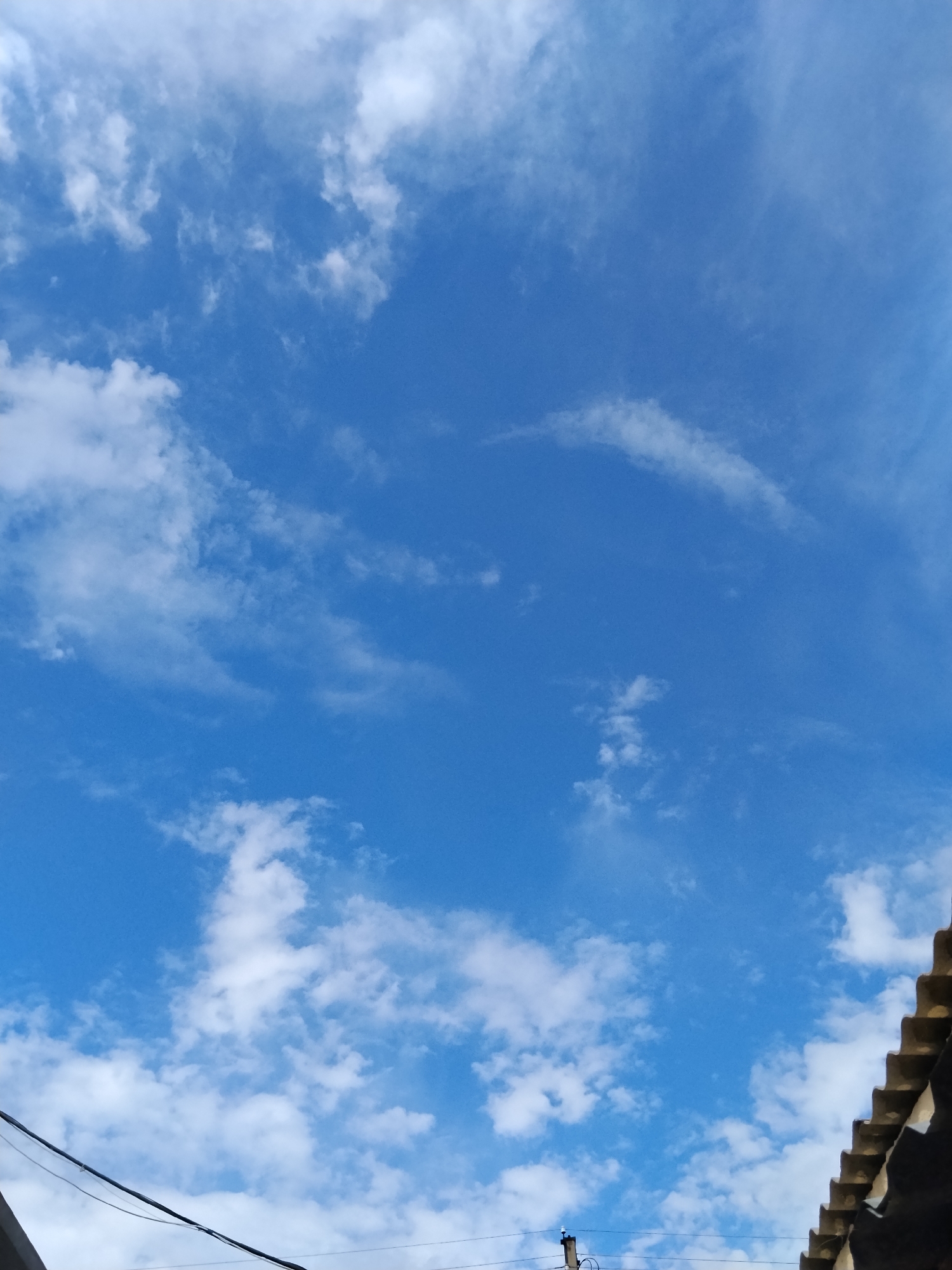 The image shows a bright blue sky with scattered white clouds. In the lower right corner, the edge of a dark, tiled roof is visible, and a black cable runs diagonally across the bottom left of the frame. The scene appears to be an outdoor shot taken during the daytime, likely in Kokshetau, Kazakhstan, based on the provided context. There are no people or specific actions depicted. The focus is on the sky and elements of a building's rooftop.