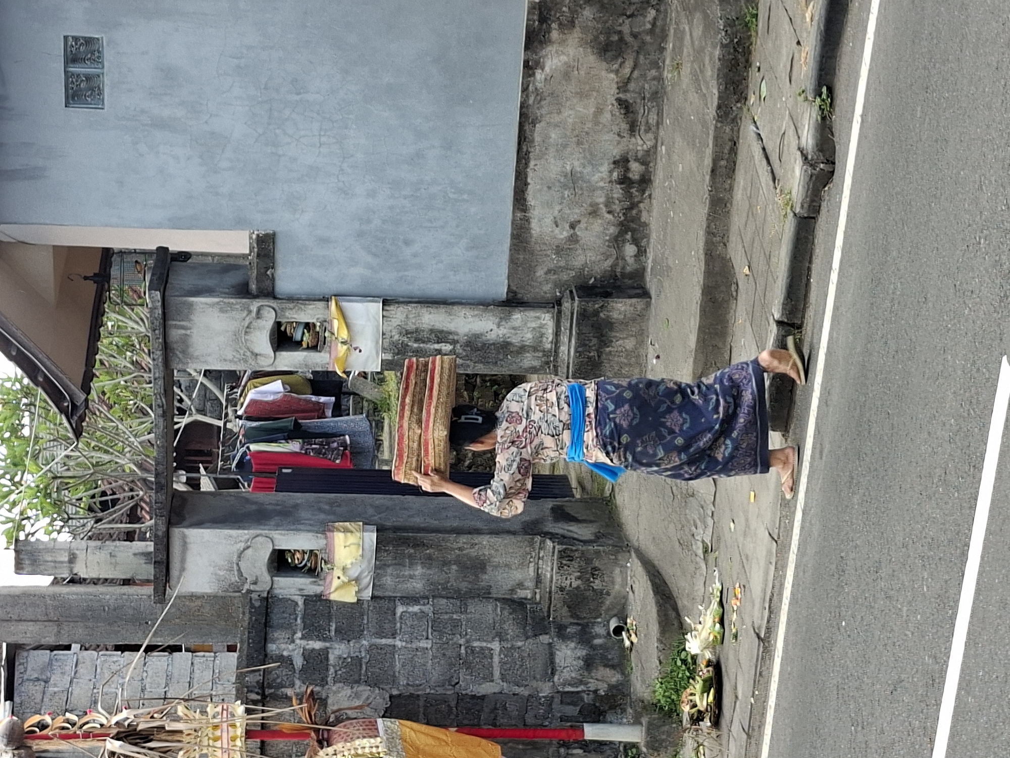 A Balinese woman, wearing a traditional patterned sarong and a light-colored patterned top with a blue sash, is walking on a sidewalk in Blahbatuh, Indonesia. She is carrying a stack of woven mats or baskets on her head. In the background, there is a weathered concrete gateway with a red and yellow offering basket on the left side, and clothing hanging on a rack through the gateway. A light blue building with a small window is visible to the right of the gateway. On the sidewalk in front of the gateway, there are remnants of offerings or trash. The street is paved with asphalt and has a white line marking the edge.