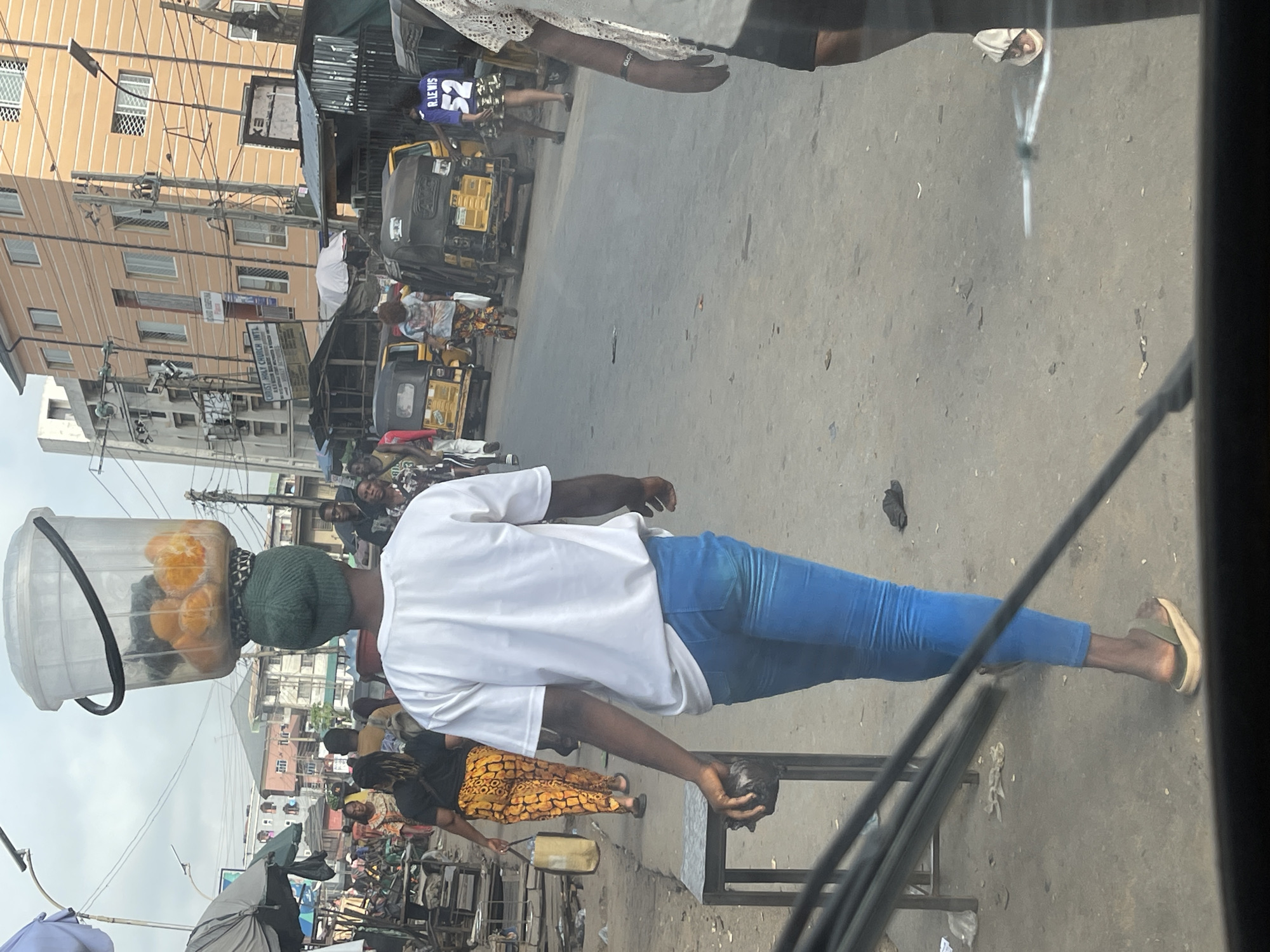 A street vendor carries a bucket of oranges on their head while walking in the city of Onitsha, Nigeria. The vendor wears a white t-shirt, blue jeans, a green knit cap, and sandals. They carry a dark object in their left hand. The street is crowded with people, vehicles, and vendor stalls. Buildings are visible on either side of the street. The picture is taken from inside a vehicle.