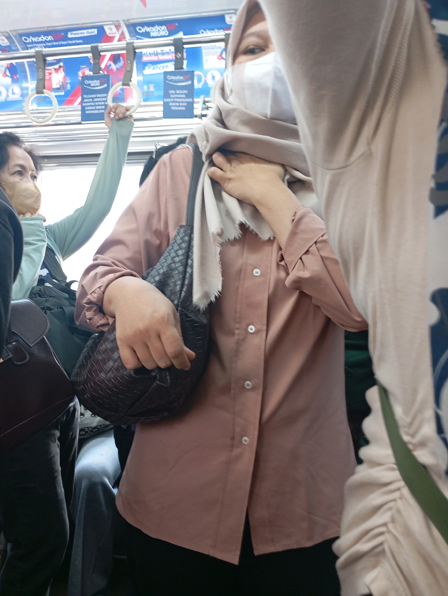 The image shows a group of people inside a public transport vehicle, likely a train or bus, in West Jakarta, Indonesia. A woman in the foreground is wearing a light brown hijab and a white face mask, obscuring her lower face. She is also wearing a pink collared shirt with buttons and a fringed scarf around her neck. She is holding a dark woven handbag with her right hand. To her left, another person is partially visible, wearing a light blue long-sleeved top and a light brown face mask. Behind them, the interior of the vehicle is visible, with metal poles and white circular handles for passengers to hold onto. Advertisements with Indonesian text are displayed above the handles. The lighting suggests it is daytime.