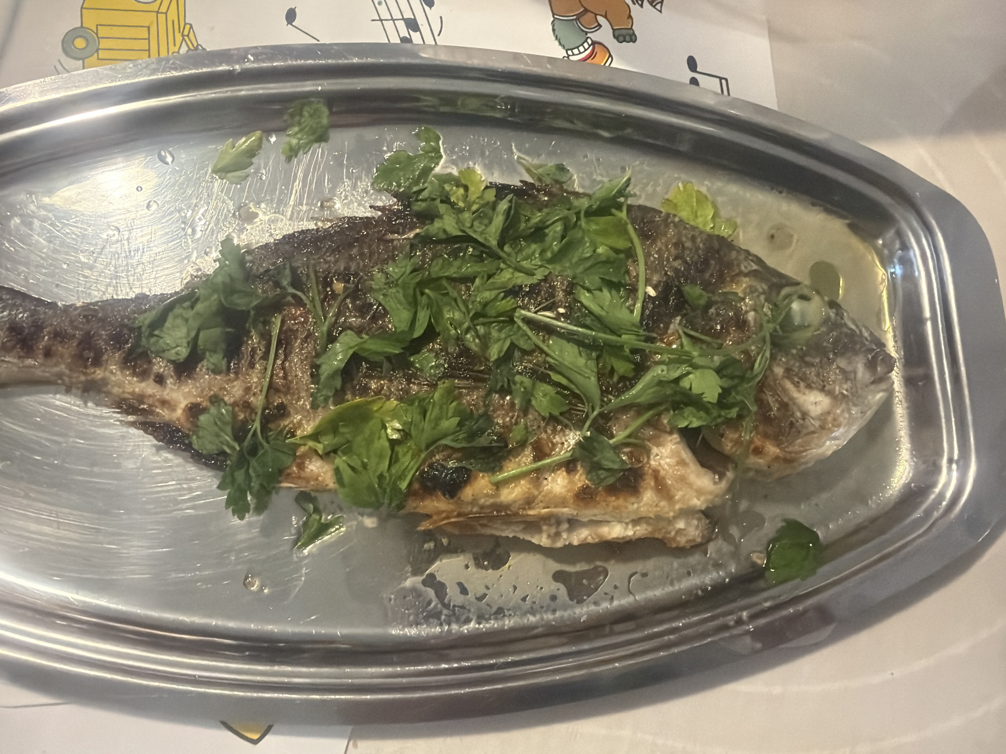 A whole roasted fish, garnished with fresh green herbs, sits on a metallic oval platter. The fish is lying on its side, its skin is slightly charred in places, indicating it has been grilled or baked. The platter is wet, with droplets of liquid pooling around the fish. The background shows a white surface with some cartoonish drawings of musical notes and what appears to be a teddy bear wearing a green sock. The platter is placed on a white tablecloth. The overall scene suggests a meal being served, possibly in a casual dining setting. The lighting appears to be indoor. No people are visible in the frame.