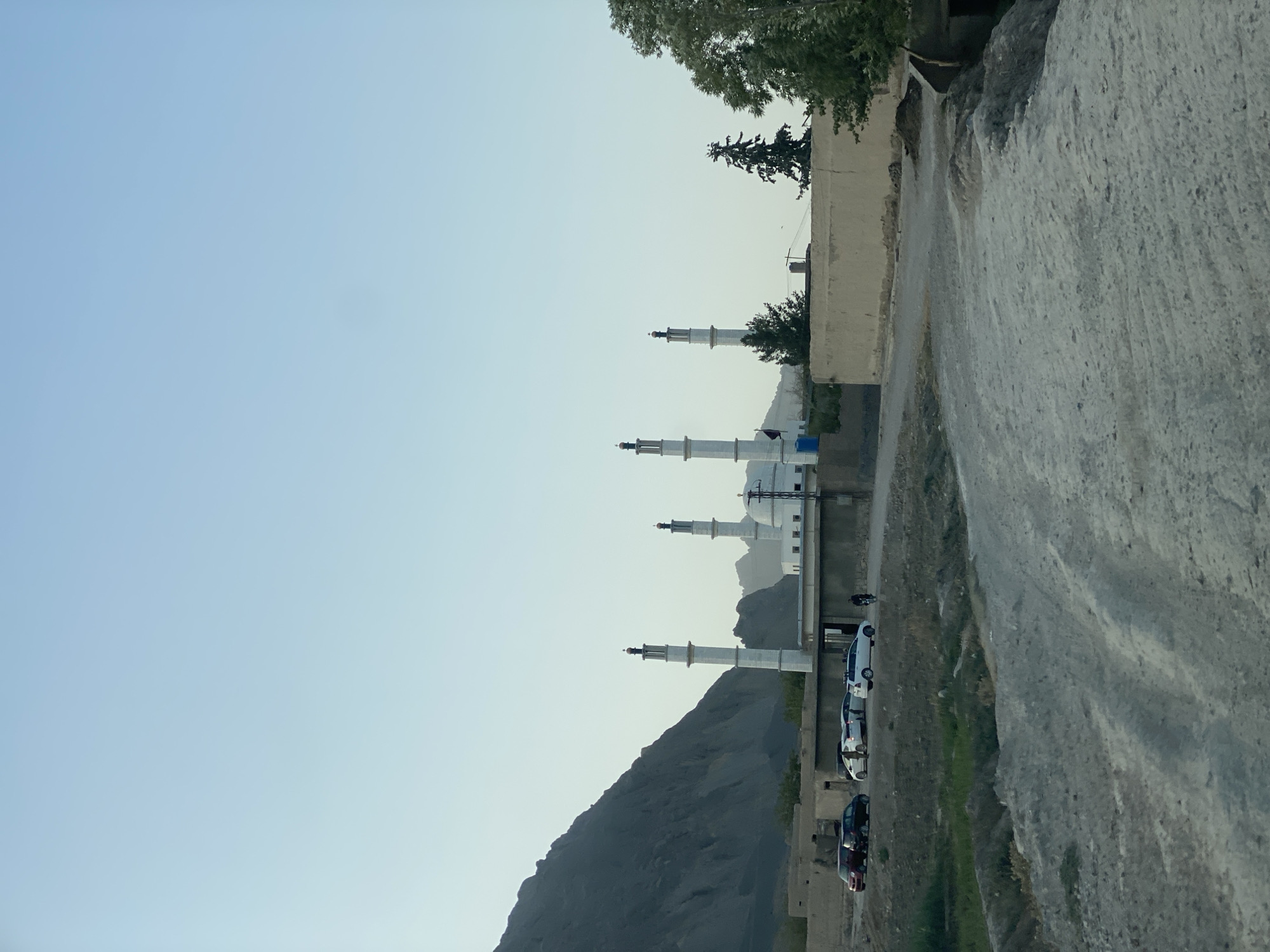 The image shows a mosque with multiple minarets and a dome, located in a relatively rural or semi-rural area of Kandahar, Afghanistan. The mosque is constructed with white minarets and a white dome, and is surrounded by a wall. In front of the mosque, there is a parking area with several cars, including a red sedan, a black sedan, a white sedan, and a white SUV. Some people are visible near the cars and entering the mosque. A dirt road or path leads towards the mosque, with dry vegetation and some green patches of grass along the sides. In the background, there are mountains, characteristic of the landscape in and around Kandahar. The sky is clear and pale blue, indicating it is likely daytime, possibly late afternoon due to the soft light. A few trees are also present, adding to the natural environment. The overall scene suggests a functioning place of worship and community gathering in the outskirts of the city.