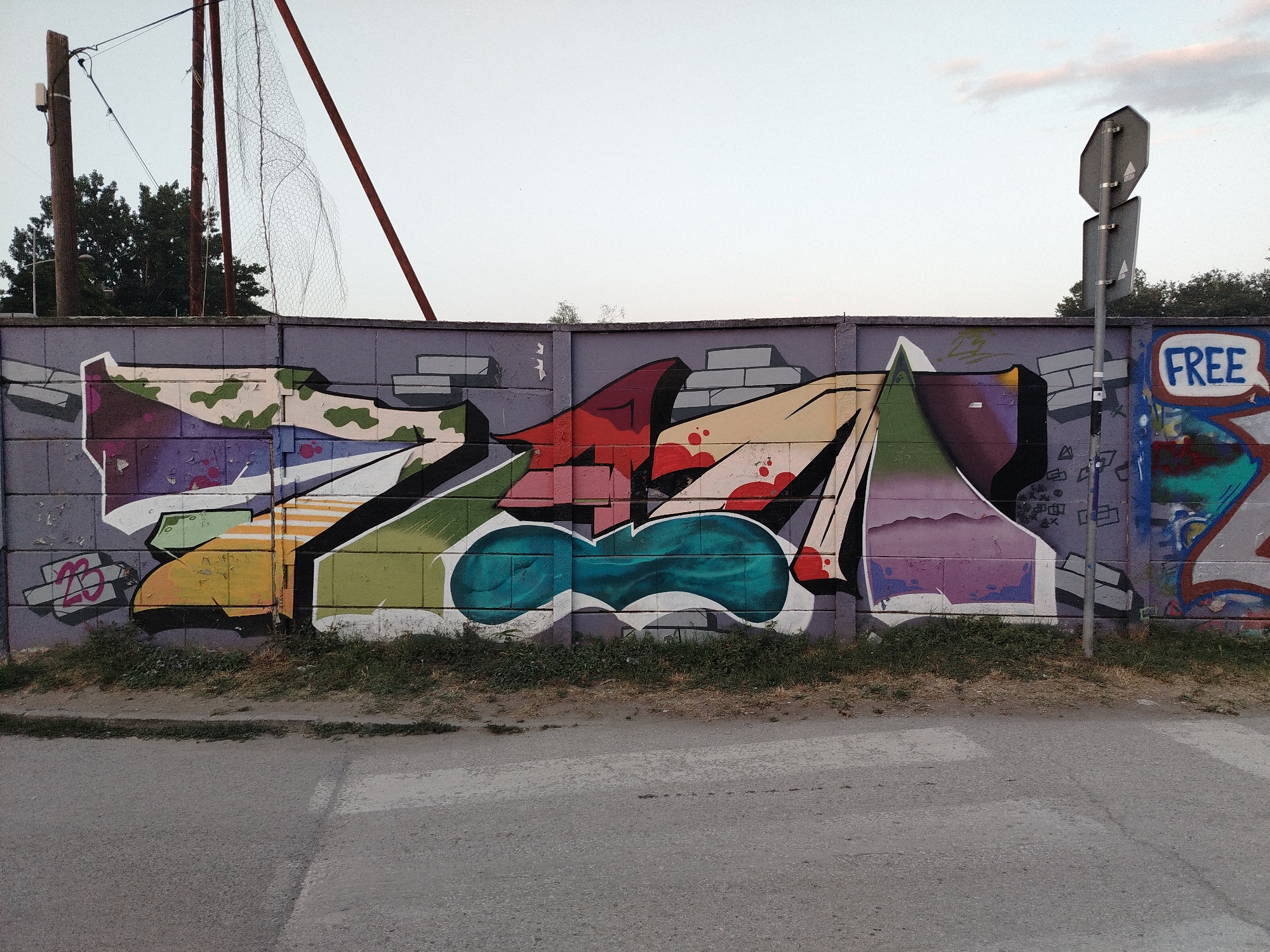 The image shows a section of a graffiti-covered concrete block wall in Novi Sad, Serbia. The graffiti features bold, colorful lettering and abstract shapes in shades of red, yellow, green, purple, and blue. To the right of the main graffiti, a speech bubble with the word "FREE" written in blue letters is visible. A metal pole with two traffic signs is positioned in front of the wall on the right side of the image. Behind the wall, trees and a sky with scattered clouds are visible. In the foreground, there is a paved road with a pedestrian crossing marked by white lines. The ground in front of the wall is covered with dry grass and some weeds. The overall scene depicts an urban environment with street art and public signage.