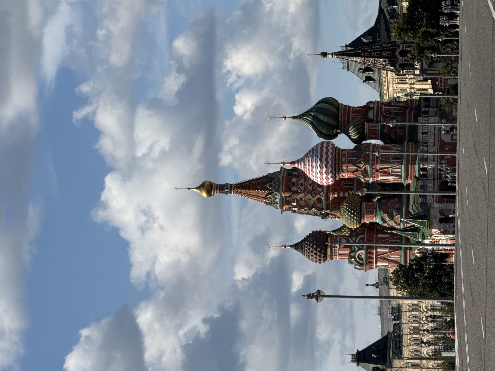 The image shows Saint Basil's Cathedral in Moscow, Russia, under a partly cloudy blue sky. The cathedral is the central subject, with its iconic colorful onion domes and intricate architectural details prominently displayed. To the left of the cathedral, a portion of the State Historical Museum is visible, characterized by its light-colored facade and dark green roof. To the right, another building with similar architectural style can be seen. The foreground features a wide, paved road with white lane markings, suggesting a public thoroughfare. Several lampposts are scattered throughout the scene. A few people are visible in the middle ground, near the buildings, and the general environment suggests an outdoor, urban setting. The overall impression is a clear day with a mix of sun and clouds.
