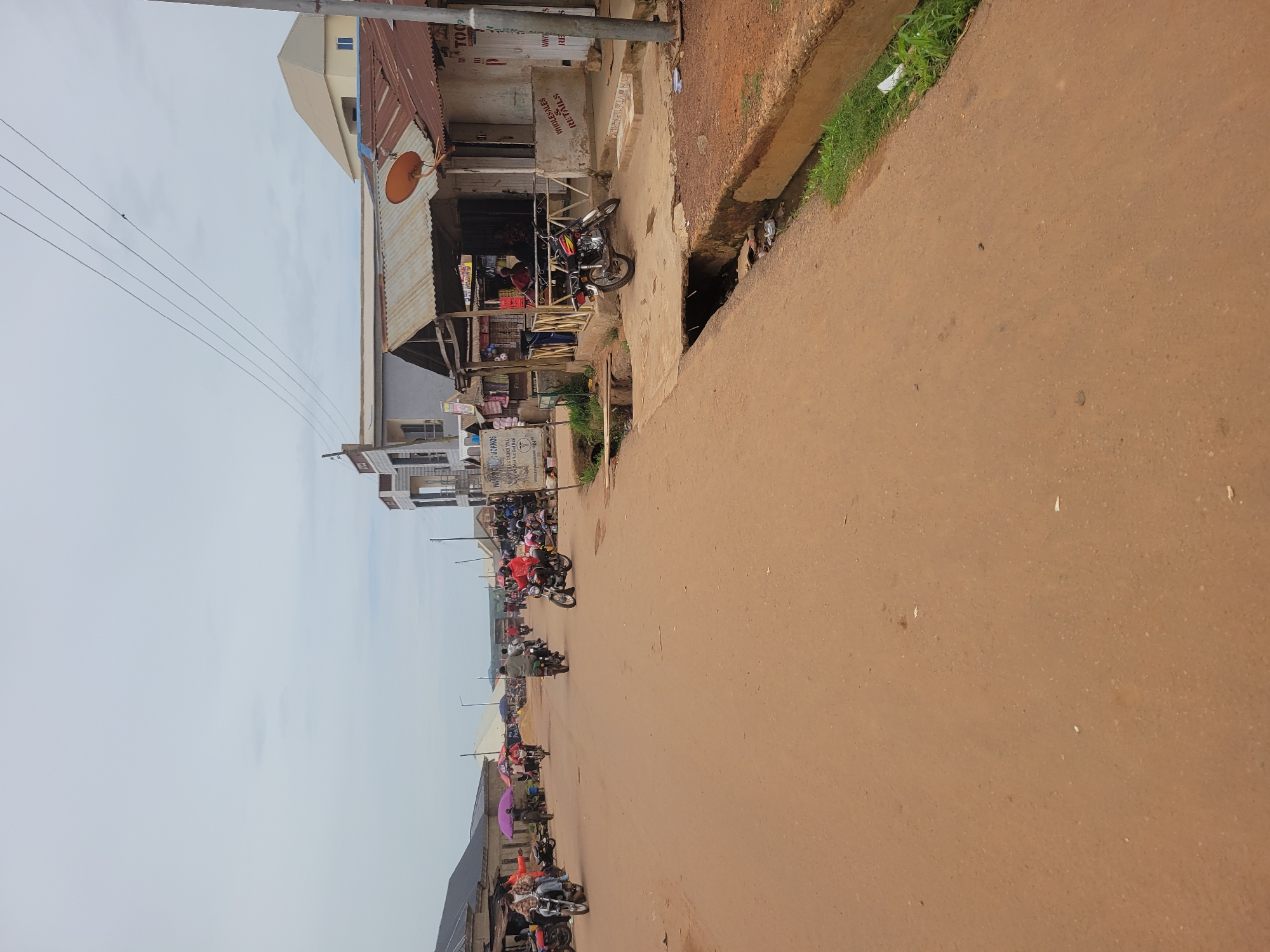 This is a photograph of a street scene in Bokkos, Nigeria, on an overcast day. The road is made of reddish-brown earth and appears to be dusty. Several motorcycles are present, some ridden by people and others parked. On the right side of the road, there are buildings with corrugated iron roofs and painted walls. One building has signage that reads "WHOLESALES & RETAILS". A satellite dish is visible on one of the roofs. A drainage channel runs along the side of the road, with some greenery growing in and around it. People are walking and standing in small groups near the buildings and on the road. The overall impression is of a busy, bustling local market or commercial area within the town.