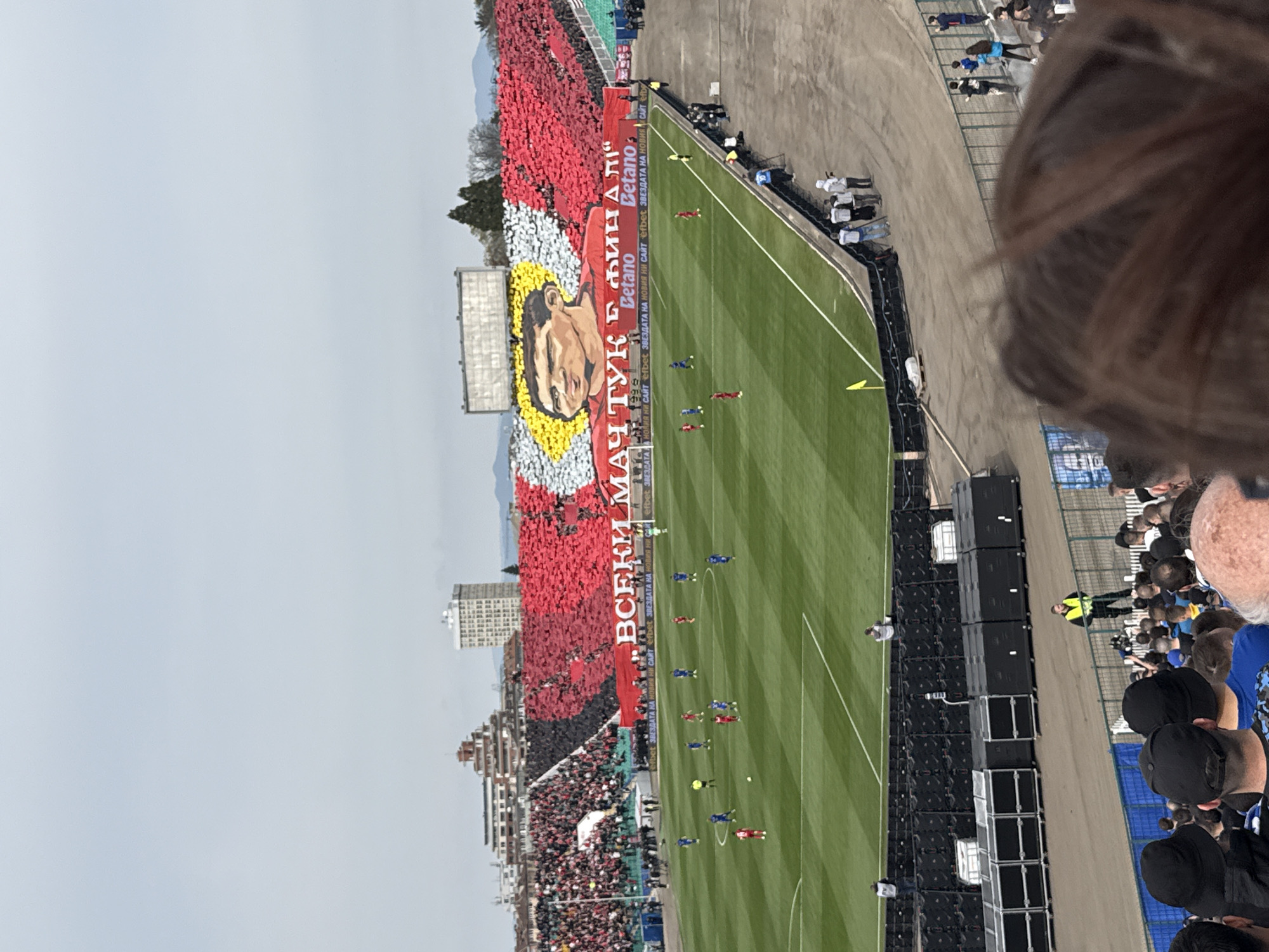 The image shows a soccer game in progress at a stadium. The field is green and there are players in blue and red uniforms. On one side of the field, a large section of the stadium is filled with spectators holding up red, yellow and white cards to create a mosaic of a man's face, along with the text "ВСЕКИ МАТУК Е ФИНАЛИ". There are advertisements for "Betano" and "efbet" visible along the side. On the other side of the field, there are speakers and more spectators, some wearing blue. The environment includes buildings visible in the background and an overcast sky. Part of a person's head is visible on the right side of the image, suggesting it was taken from the stands. The location is in an urban setting.
