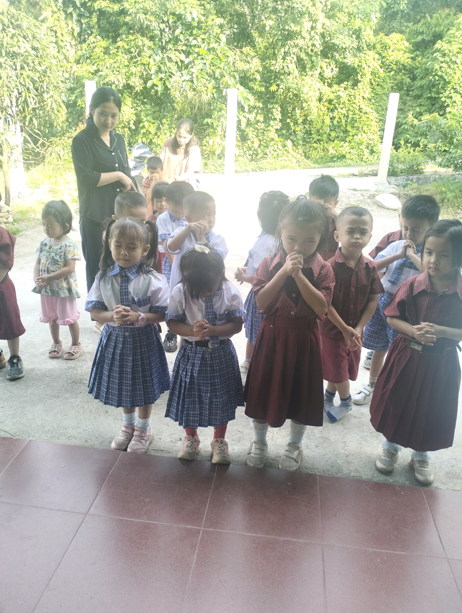 In Bonia Hilisimaetano, Indonesia, a group of children in uniform stand in a line, some with hands clasped. A woman in a black top stands behind them, and another woman in a light brown shirt is further back. The children appear to be of primary school age, dressed in various school uniforms consisting of plaid skirts and collared shirts or blouses. Some children have their eyes closed, suggesting they are praying or listening intently. The setting appears to be outdoors, with lush green foliage in the background and a tiled floor in the foreground. The scene suggests an educational or communal gathering.