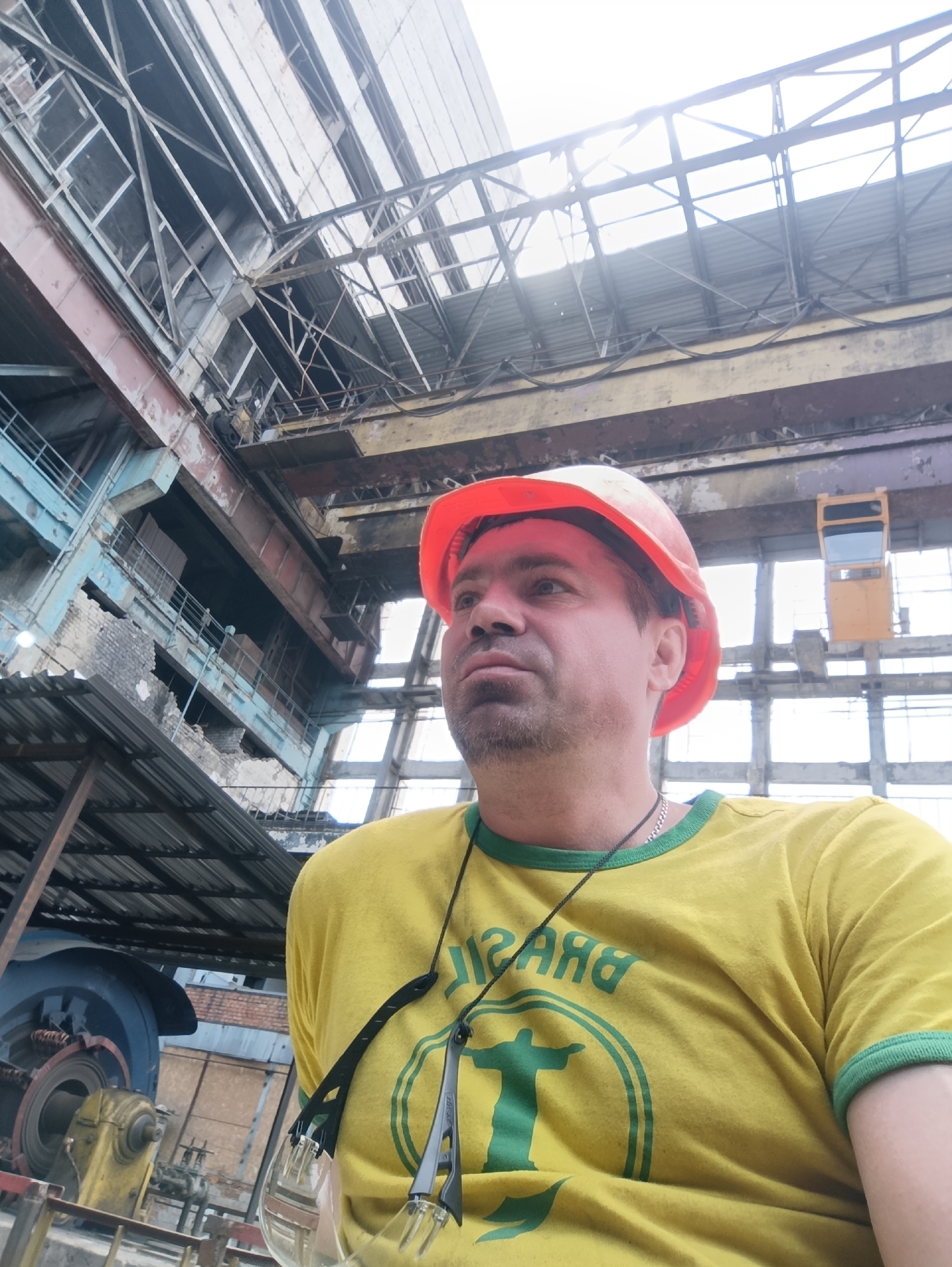 A man wearing a yellow t-shirt with a green trim and a Brazil national team logo, along with an orange hard hat, is positioned in the foreground, looking to the right. He has a beard and a silver chain around his neck. Safety glasses are hanging from his neck. The background features the interior of an industrial building, likely an industrial or power plant, in Ladyzhyn, Ukraine. The structure includes exposed metal beams, a crane, scaffolding, and large machinery, suggesting an active or undergoing renovation site. The lighting suggests an overcast day with sunlight filtering through the roof.