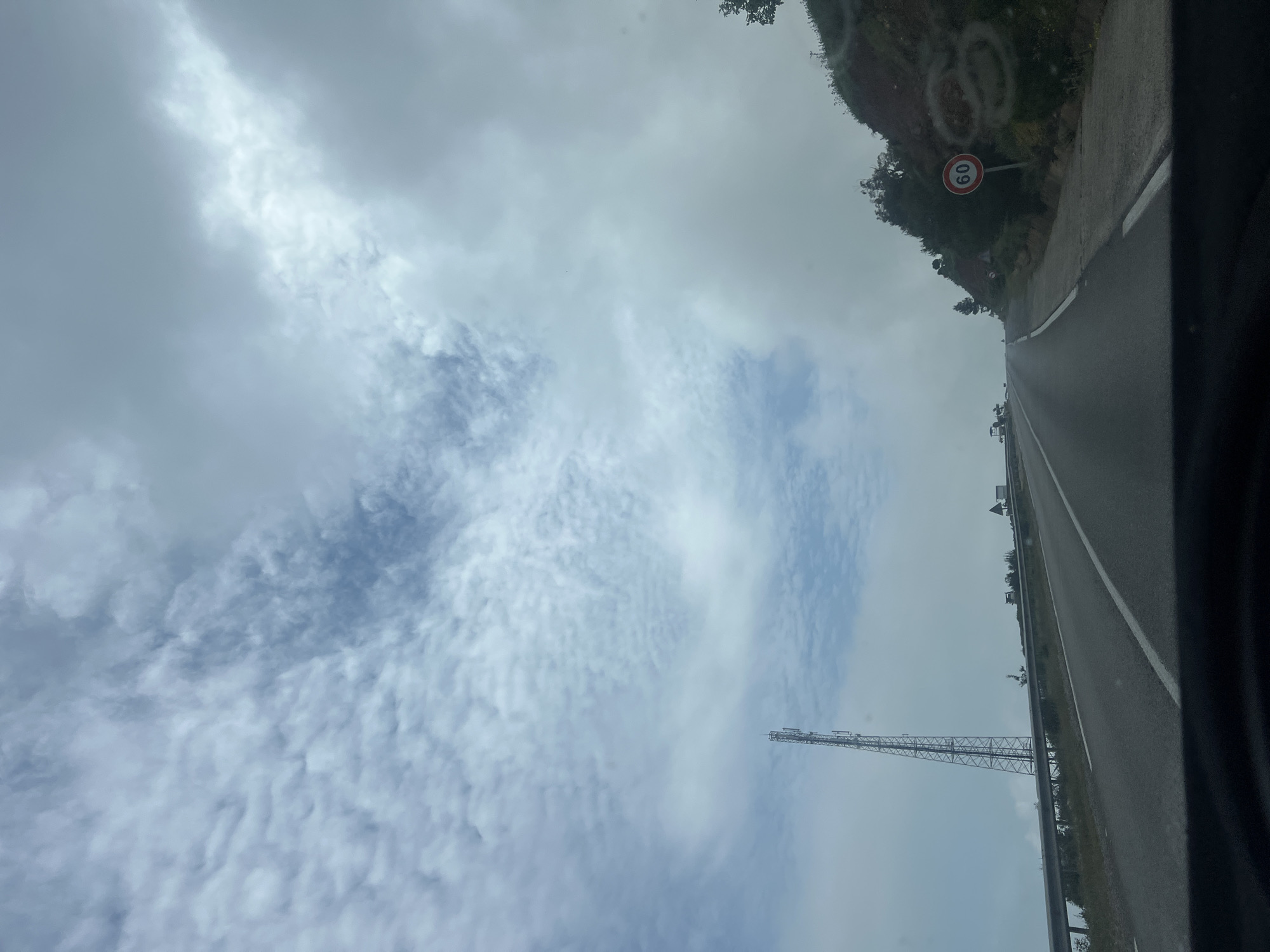 The image depicts a road in Zaouiat Sidi Kacem, Morocco, possibly taken from a moving vehicle. The road is asphalt with a white line dividing lanes. A speed limit sign (60) is visible on the side of the road. In the distance, there is a communications tower. The sky is overcast with textured clouds. The view is slightly tilted. The overall scene is a highway or major road passing through a hilly or mountainous area.