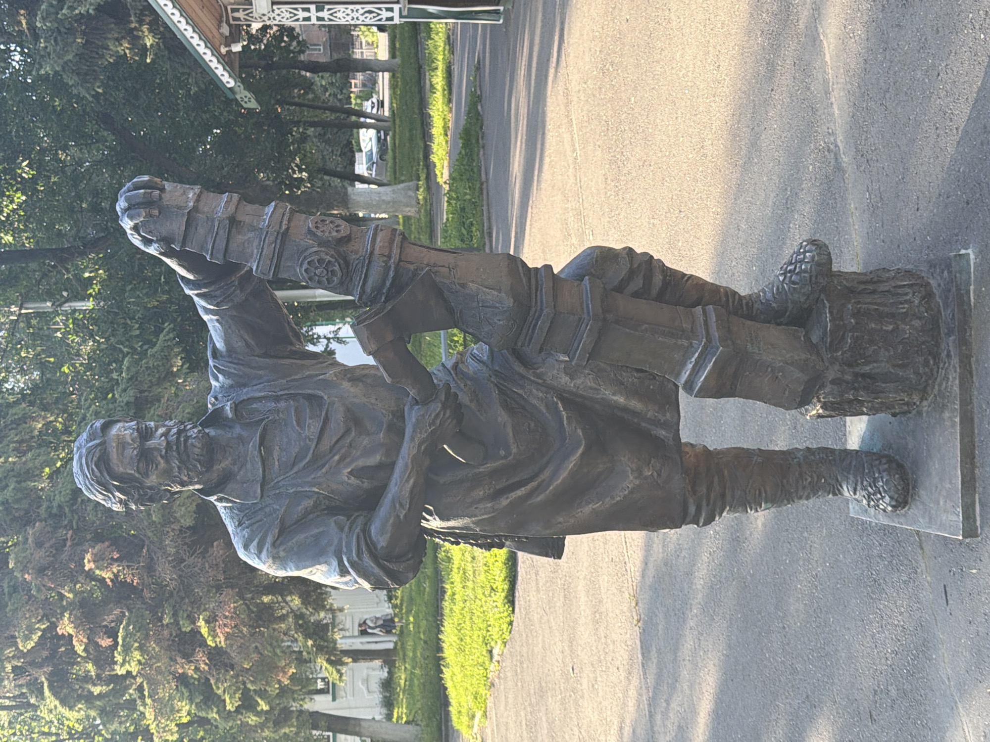 A bronze statue of a man stands on a paved surface in front of a grassy area and trees. The man is depicted in work clothes, including a tunic, apron, and tall boots, and he is holding onto a large, ornate post with both hands. His beard and mustache are prominent, and his gaze appears to be directed forward. The statue is set on a square base, which is resting on a larger rectangular plinth. In the background, trees, a white building with decorative trim, and cars on a road are visible. The overall lighting suggests it is daytime with sunlight casting shadows. This statue is located in the city of Vologda, Russia, and depicts a locksmith.