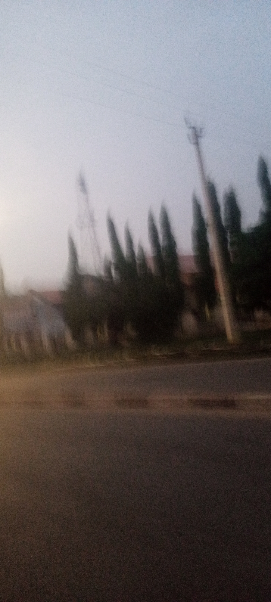 The image depicts a blurred urban scene at twilight in an unknown location. A paved road is in the foreground. Beyond the road, there's a row of tall, slender trees. A building is visible behind the trees, and an electrical pylon stands in the background. An electrical pole is also seen near the trees on the right side of the image.