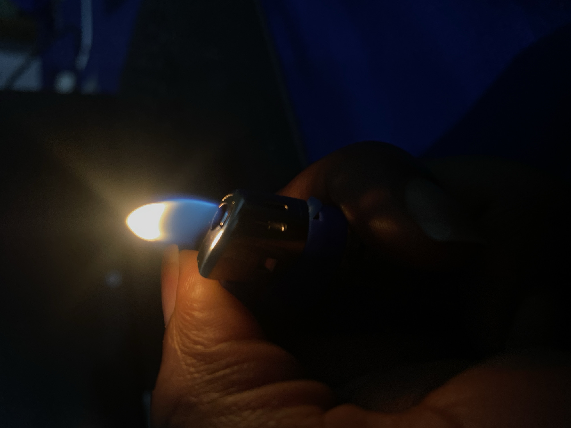 A hand holds a lit blue lighter in a dark environment. The flame is yellow at the base and blue at the tip. Details of the hand are obscured by shadow. The background is dark, with some vague shapes suggesting objects, including something blue. There is no visual information to determine if this is Jos, Nigeria.