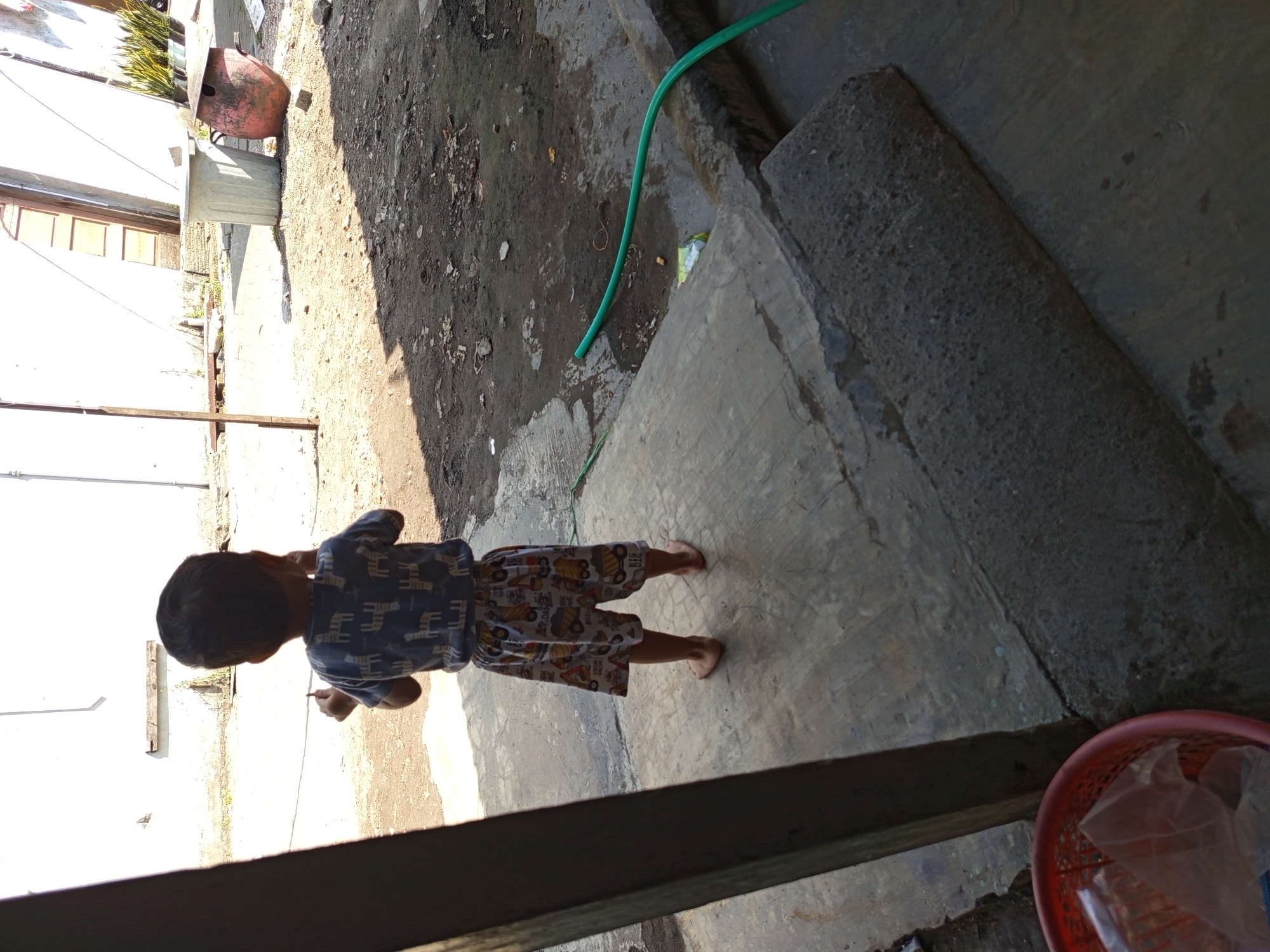 A child stands barefoot on a concrete patio, facing away from the camera. The child is wearing a blue t-shirt with a white pattern and patterned shorts featuring construction vehicles. Sunlight casts a strong shadow from a wooden beam on the left side of the image. The background reveals a dirt and debris-strewn yard with a white wall, a grey trash can, and a red bucket with plastic bags inside. A green garden hose snakes across the ground in the middle ground. The scene appears to be an outdoor area of a residential property in Tebel, Indonesia.