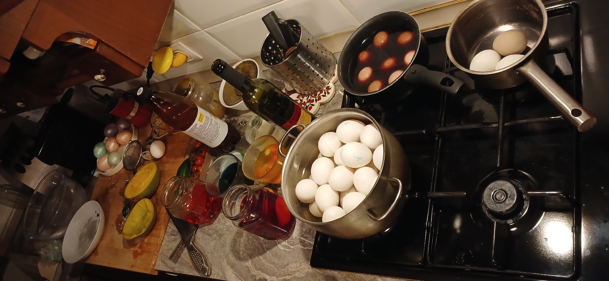 The image captures a kitchen scene during what appears to be Easter egg preparation, at an unidentified location.
Environment: The kitchen features white tiled walls and dark countertops. A wooden cutting board is visible on the left side.
Objects:
* Eggs: A large pot overflowing with white eggs sits on a black stovetop. Some eggs are also visible in a smaller pot on the right, and another pot containing colored eggs is located near the center. On the left, a plate displays a variety of dyed eggs.
* Liquids: Several glass jars containing colored liquids, presumably for dyeing eggs, are arranged on the counter. Bottles of liquids, possibly ingredients for dyeing or other kitchen items, are also present.
* Utensils: A metal strainer and other kitchen utensils are scattered around the countertop.
* Fruits: Cut pieces of yellow fruit are visible on the cutting board.
* Other: The stovetop is black and clean. A metal cutlery holder sits next to the egg-dyeing setup.
Actions: The scene indicates egg preparation. Eggs are being boiled, dyed, and possibly sorted.
Interactions: There's no visible interaction with people. The scene suggests the process of dyeing eggs is actively underway or has recently concluded.