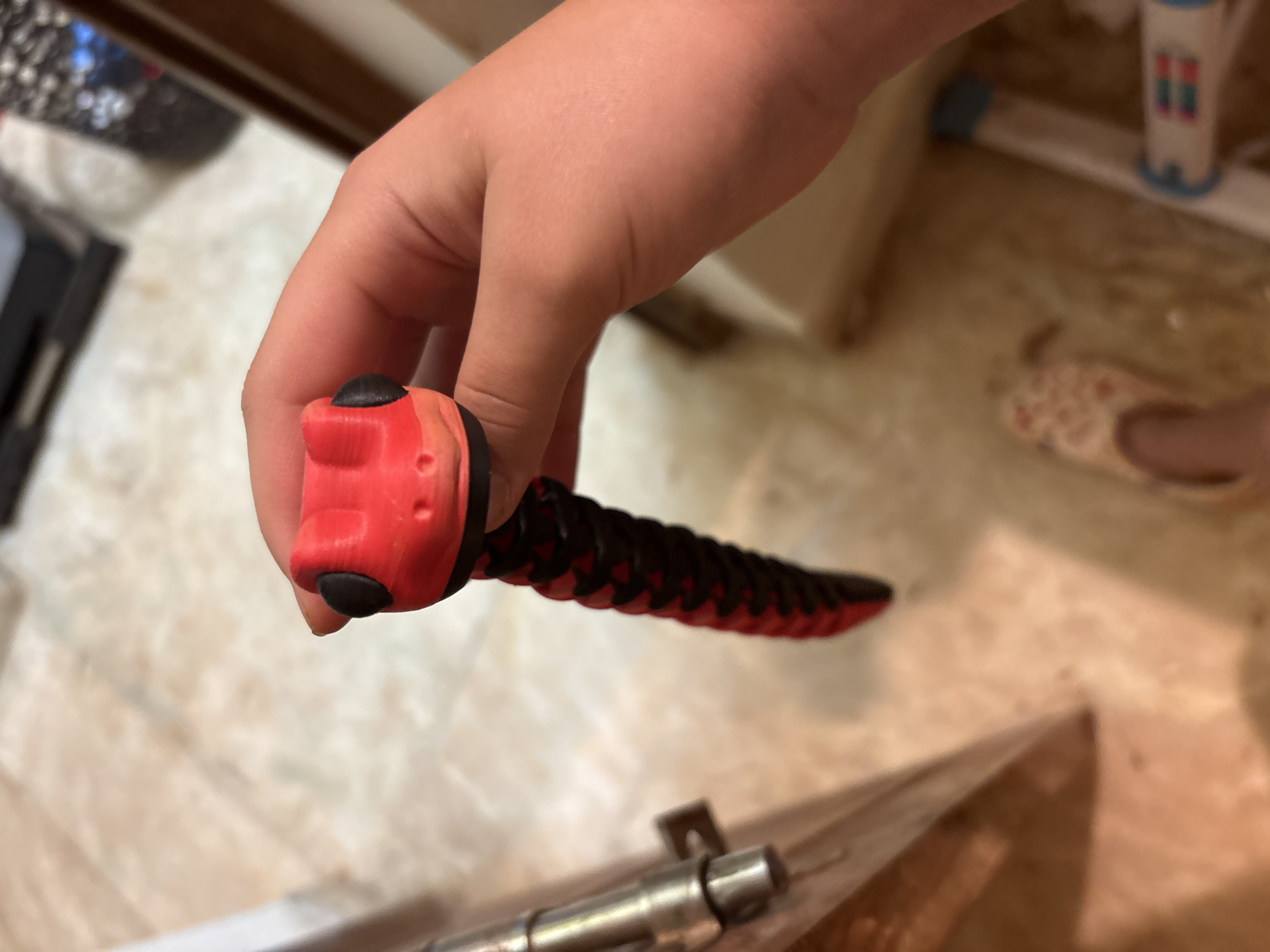 A close-up, slightly angled shot shows a person's hand holding a red and black segmented toy snake. The snake's head is red with black circular eyes and a small mouth. Its body is segmented, alternating between red and black sections. The background is blurred and appears to be an indoor setting with light-colored flooring and a hint of wooden furniture on the left. A glimpse of a shoe with a floral pattern is visible near the bottom of the frame. The image does not provide any information about the city of Ashvem, India.