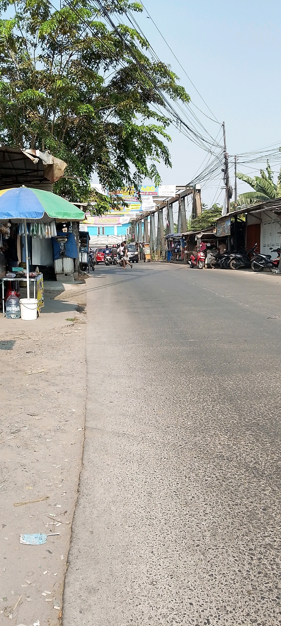 This is a ground-level, eye-level shot of a street scene in Babelan, Indonesia. The image features a paved road with traffic, including cars and motorcycles. On the left side of the road, there's a sidewalk area with a small vendor stall covered by a colorful umbrella. This stall has items displayed for sale, including bottled water and buckets. Beyond the stall, there are buildings and trees. On the right side of the road, a series of small shops or stalls line the street, with motorcycles parked in front of them. A pedestrian bridge or overpass is visible in the background, with signage on it. The sky is clear and blue. The overall impression is of a bustling local street environment.