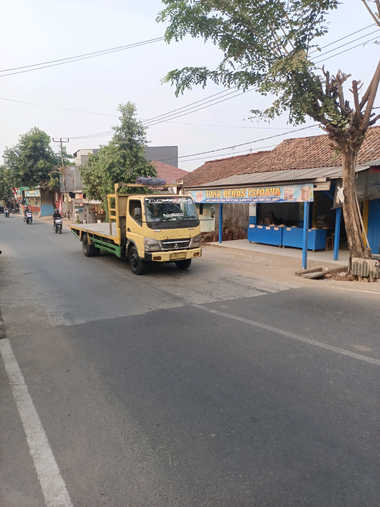 A yellow flatbed truck is driving down a two-lane asphalt road in Karawang, Indonesia. The truck has a wooden flatbed with green metal sides and is carrying a large green tree on its bed. The truck's front is facing the right side of the image. On the right side of the road, there is a building with a red tile roof and a blue awning that reads "TOKO BERAS ISMAYA." There are several blue display cases under the awning. On the left side of the road, there are trees and buildings, and several motorcycles are visible. The sky is a pale blue with some power lines overhead. The scene appears to be a street in a moderately populated area.