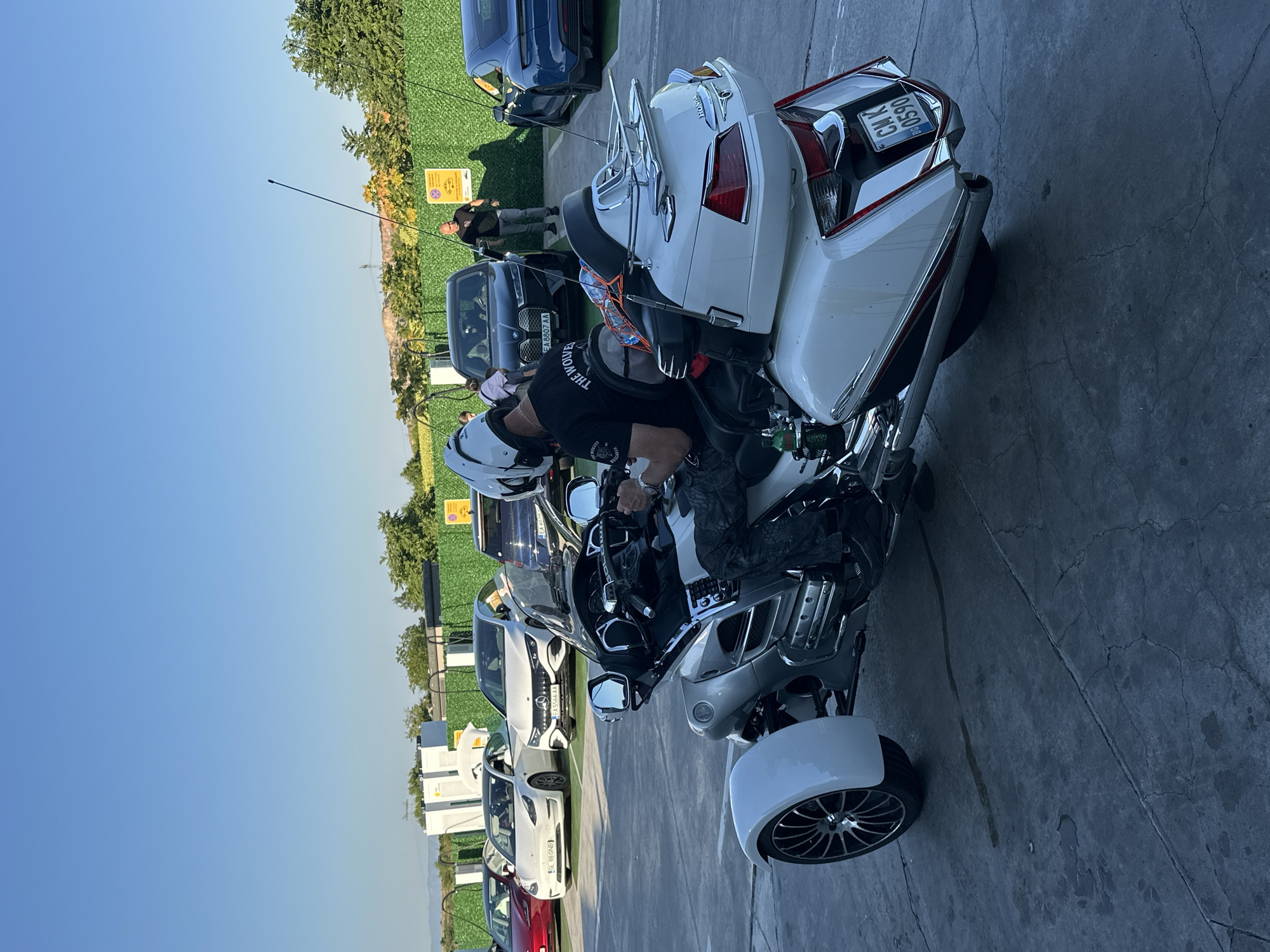 In Polsko Padarevo, Bulgaria, a white three-wheeled motorcycle, possibly a Honda Gold Wing, is parked on a cracked asphalt surface, likely at an electric vehicle charging station, indicated by the charging stations in the background. The motorcycle is facing to the right, with its rear prominently displayed. A person wearing a white helmet and a black t-shirt with "THE WOLVES" written on it is seated on the motorcycle, holding the handlebars. In the background, several cars are parked, including a white Tesla, a white Mercedes-Benz, and a dark blue Tesla. A man in a black t-shirt is standing near the dark blue Tesla, and other individuals are visible in the distance. The environment appears to be an outdoor area with a clear blue sky and some greenery. The license plate on the motorcycle reads "CM K BG 0590".