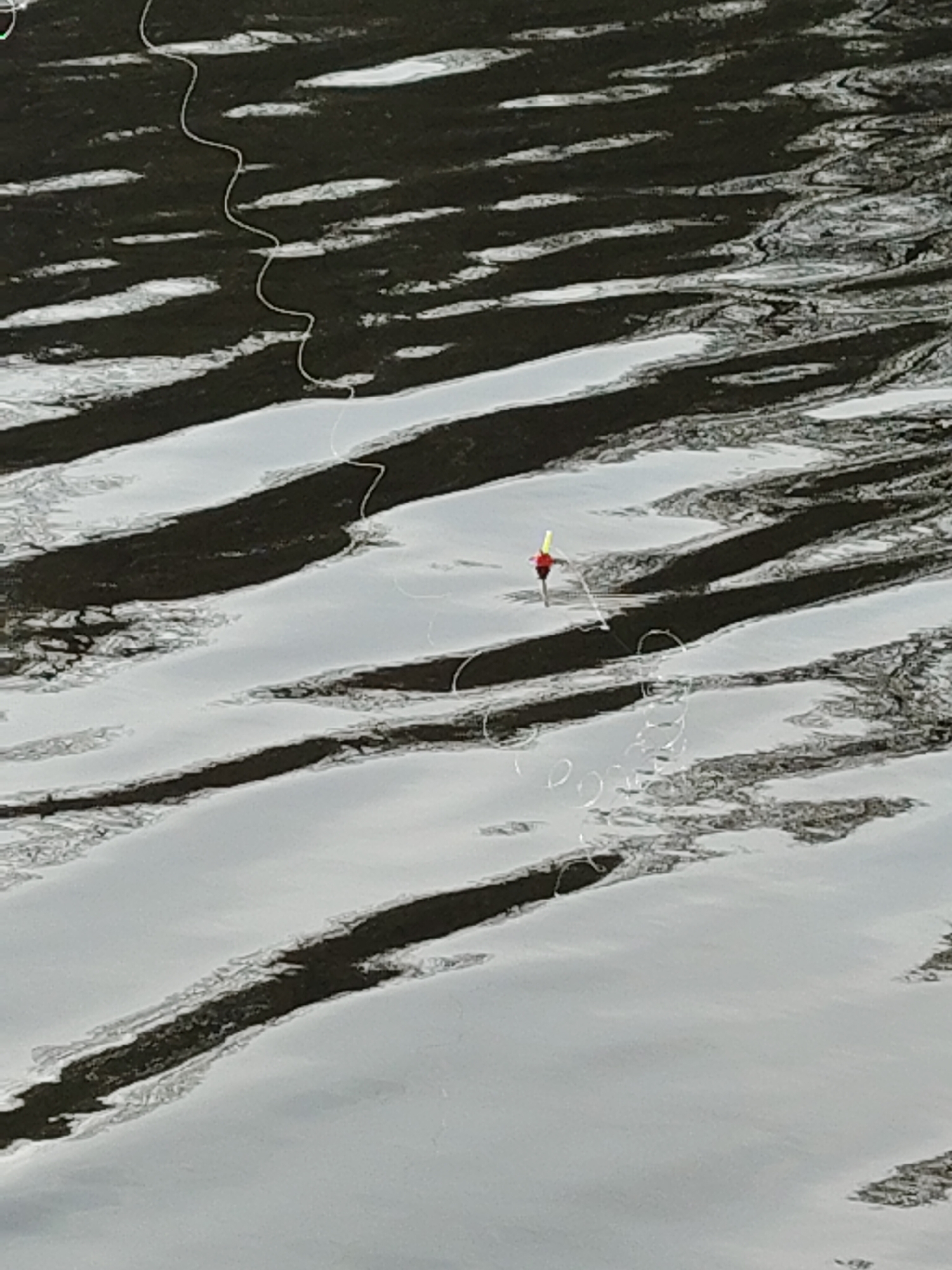 The image depicts a body of water with ice formations and a fishing bobber. The water surface has horizontal stripes created by the interplay of light and shadow on the ice. A red and yellow fishing bobber floats on the water, connected to a fishing line that extends out of frame. The location is Unknown, Unknown.