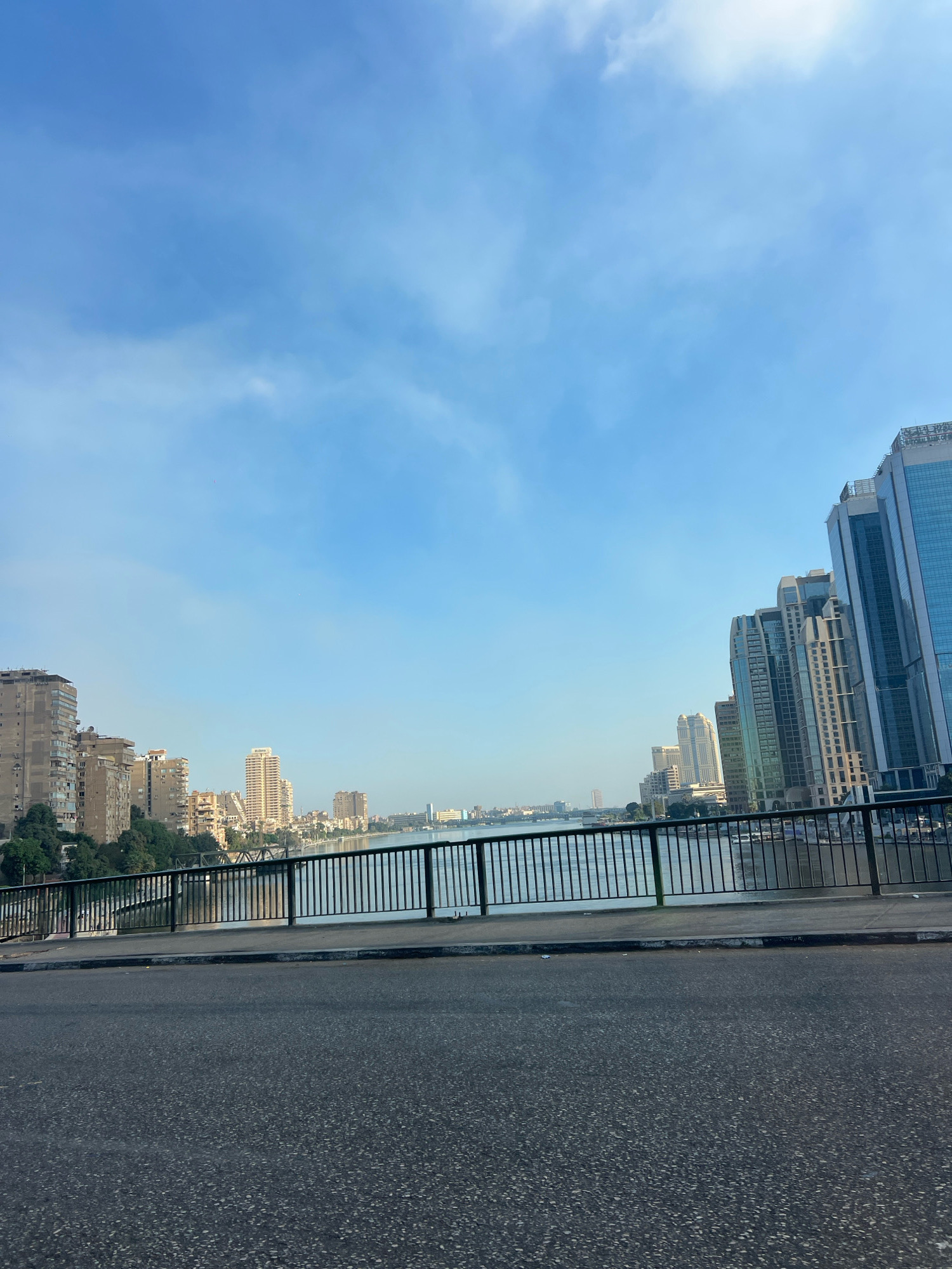 The image captures a view across a river in Cairo, Egypt, likely the Nile River, from a bridge. On the left side of the frame, several mid-rise apartment buildings are visible, interspersed with trees. As the view moves to the right, a modern skyline of tall, glass-facade skyscrapers dominates the opposite bank. A black metal railing runs along the edge of the bridge in the foreground, separating the walkway from the river below. The river itself is calm, reflecting the light blue sky with scattered white clouds. The road surface of the bridge is visible in the lower portion of the image, appearing dark and asphalted. The overall scene suggests a city view during daytime, possibly morning or late afternoon, given the quality of light. There are no people or vehicles immediately discernible in the frame.